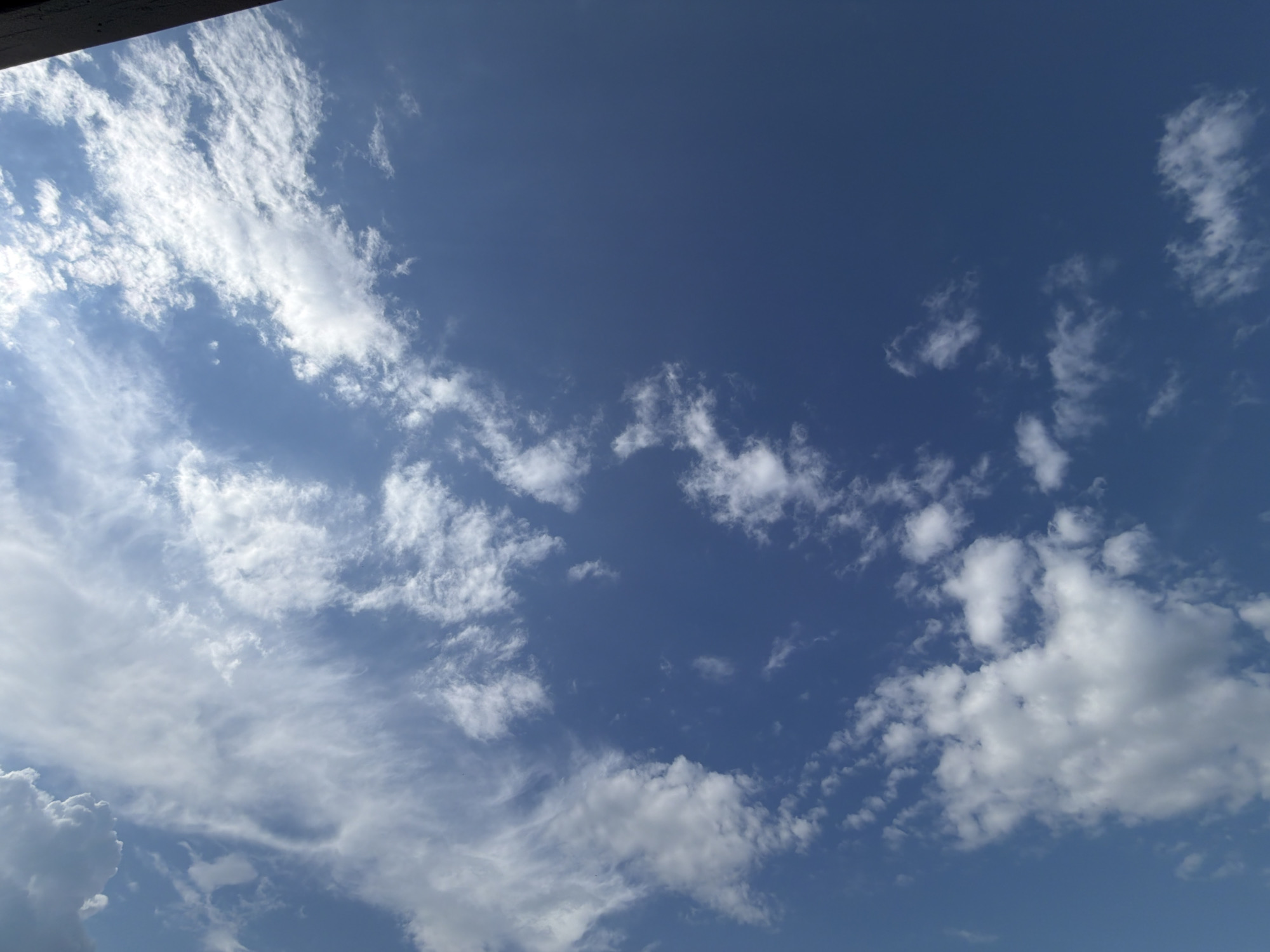 The image shows a sky with scattered white clouds against a deep blue background. The clouds vary in density and shape, with some appearing wispy and others more voluminous. The lighting suggests it is daytime, possibly with the sun partially obscured or diffused by the clouds. The upper right corner of the image features a dark, vertical rectangular object, likely a building or structure, casting a shadow or simply being part of the foreground. No people or specific city landmarks are visible to identify the location.