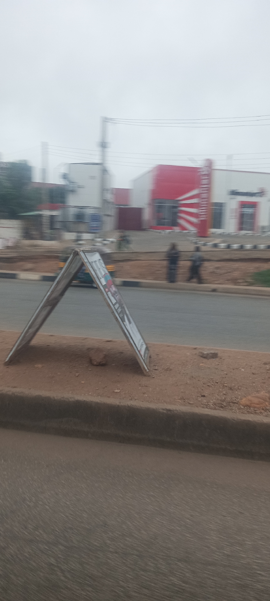This slightly blurred, outdoor photograph depicts a street scene in Jos, Nigeria, under an overcast sky. In the foreground, a white A-frame sign stands on a raised dirt median, its contents unreadable due to blur and distance. The median is edged by a concrete curb alongside a paved road.
Across the road, two indistinct human figures are visible, possibly walking or standing, near another striped curb. The background features several buildings. The most prominent structure on the right is a white building with a large, distinctive red section on its facade, adorned with white radiating lines. This design strongly resembles the logo of KFC, and faint, vertically oriented text consistent with "KFC" can be discerned on the red portion. Another building, white and grey, is situated to its left. Power lines and utility poles are also visible against the diffused light of the cloudy sky. A yellow and black vehicle, possibly a tricycle, is vaguely seen on the road behind the A-frame sign. The image captures a moment of daily street life, characterized by pedestrian and vehicle movement.
