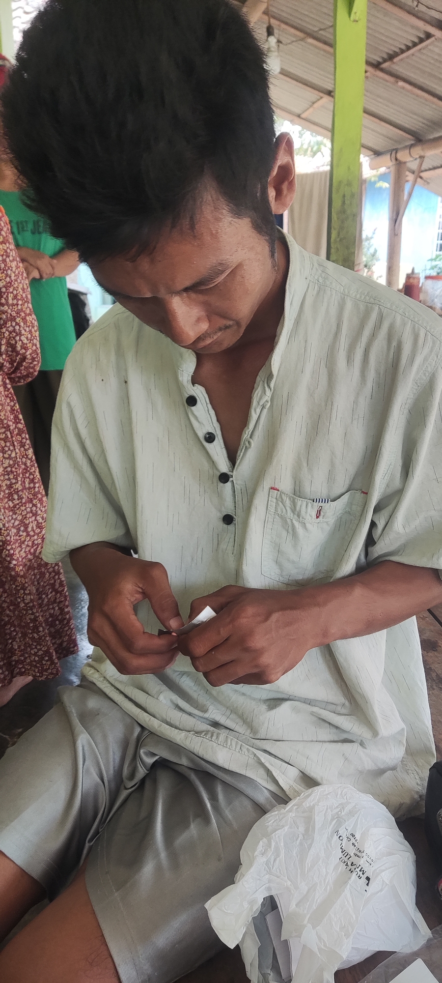 In Karangwangun, Indonesia, a person of Southeast Asian descent, with dark hair and medium-toned skin, is shown from the chest up, seated. The individual is wearing a light-colored, collared shirt with vertical dark pinstripes and black buttons. Their hands are engaged in manipulating a small, white, rectangular object, possibly paper or a packet. The background suggests an outdoor or semi-open structure, with a corrugated roof and supporting beams, one of which is painted green. Other individuals are partially visible in the background, one wearing a green t-shirt and another a patterned dress. A crumpled white plastic bag with some text printed on it is visible in the lower right portion of the frame. The overall scene appears to be a casual gathering or activity in a rural or semi-rural setting.