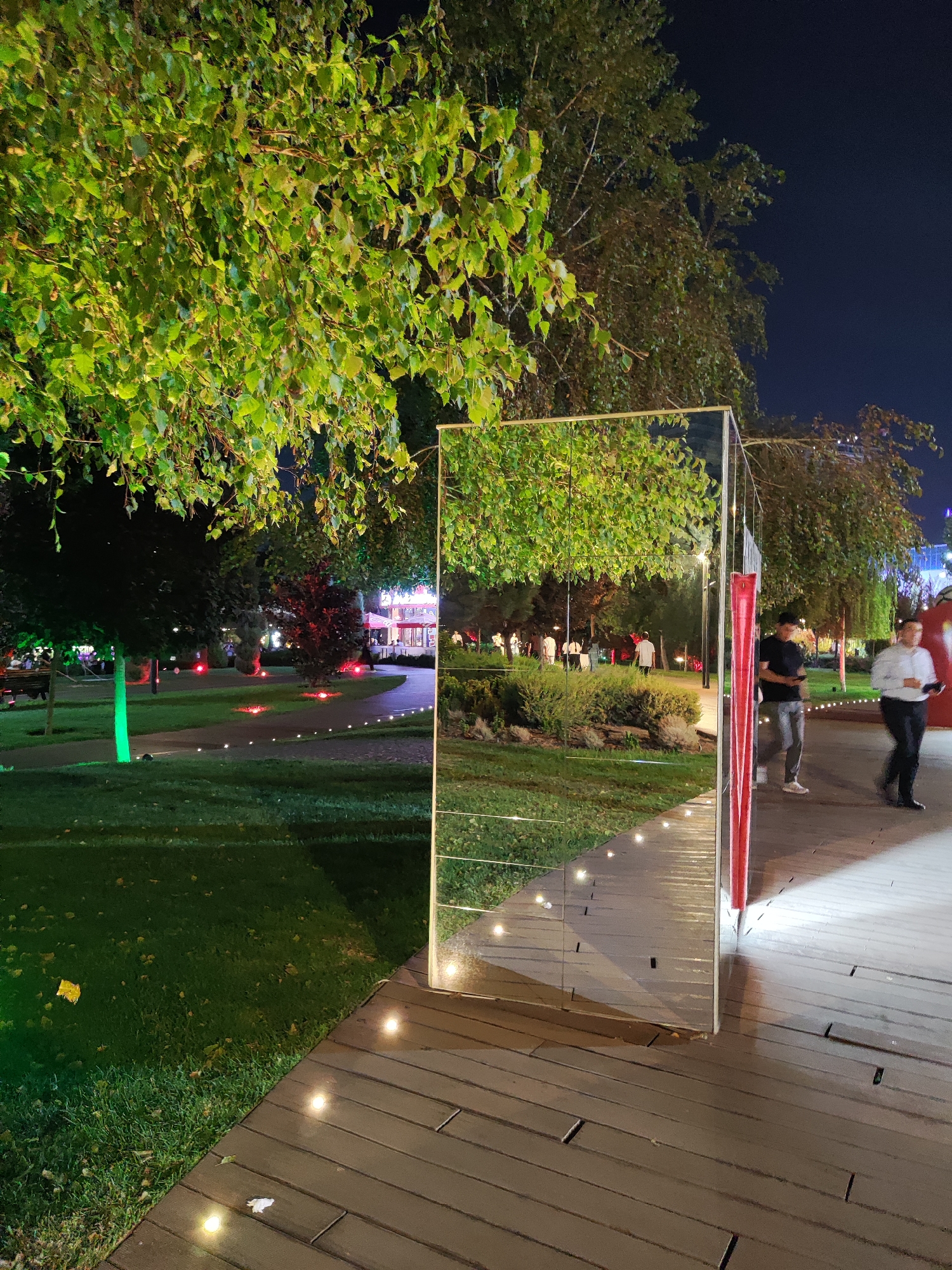 This is a night-time outdoor scene in Tashkent, Uzbekistan, likely in a park or public square. A large, rectangular mirrored art installation is positioned in the foreground, reflecting the surrounding environment. The mirror is framed by a white metal structure and a red vertical element.
To the left of the mirror, a grassy area is illuminated by green lights, with trees and foliage overhead. A pathway is visible behind the grass, also lit with small, scattered lights. In the background, there are more trees and some structures that appear to be stalls or small buildings with illuminated signs.
To the right of the mirror, a wooden boardwalk is visible, also lined with small lights. Three individuals are walking on the boardwalk. One person in a black t-shirt and grey pants is standing near the mirror. Another person in a white shirt and black pants is walking away from the camera, and a third person in a dark shirt and black pants is walking towards the camera, slightly out of focus. The sky is dark, indicating nighttime. The overall impression is a lively, illuminated public space during an evening event or gathering.