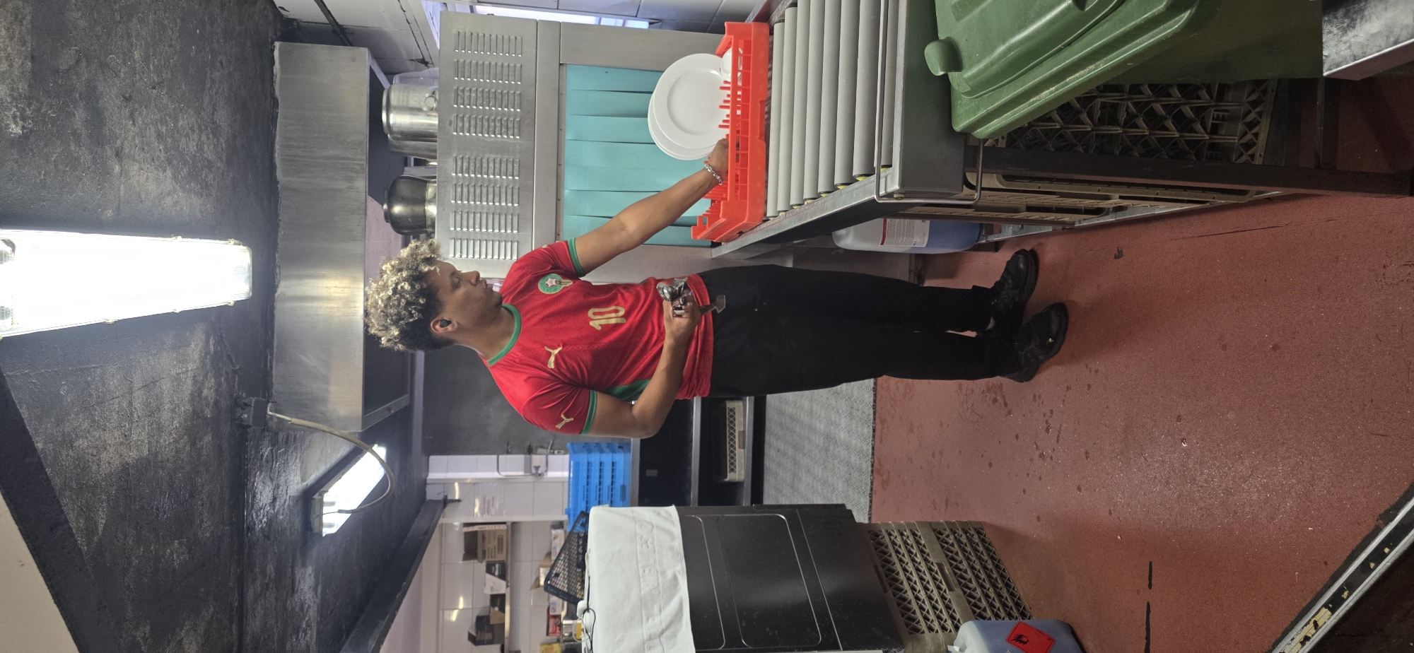 In the commercial kitchen of a restaurant in Sallent de Gállego, Spain, a young man with curly blonde hair is engaged in dishwashing. He is wearing a red t-shirt with a green trim and the number 10 on it, along with black pants and black athletic shoes. The man is holding a handheld device and is in the process of placing clean white plates into an orange dish rack. The dish rack is positioned on a conveyor belt system, which appears to be part of a commercial dishwasher. In the background, there are stainless steel appliances, blue plastic crates, and shelves stocked with various items. The floor is a reddish-brown color and shows signs of moisture. To the right of the man, a green trash bin is visible. The lighting in the scene is provided by fluorescent lights on the ceiling.