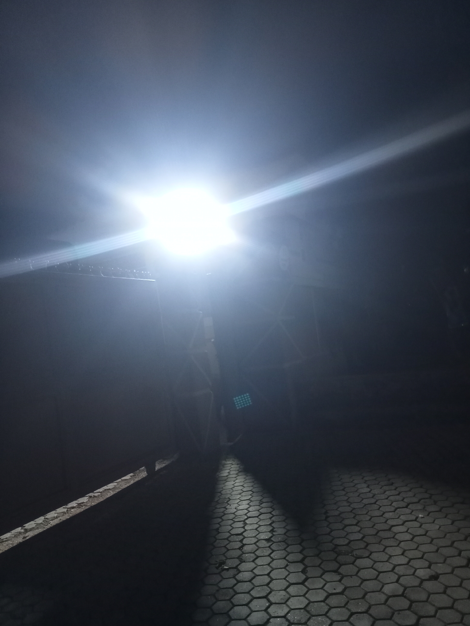 A bright, overhead light illuminates a dark scene. The light source is in the upper portion of the image, creating a strong glare and lens flare that extends diagonally across the frame. Below the light, a large, dark structure, possibly a gate or a building facade, is visible. The structure appears to have some geometric detailing. To the right and in the foreground, a paved area with hexagonal interlocking stones is visible, reflecting the light. Shadows are cast across this paved area, indicating the direction of the light. The overall impression is of an outdoor, possibly urban, setting at night.