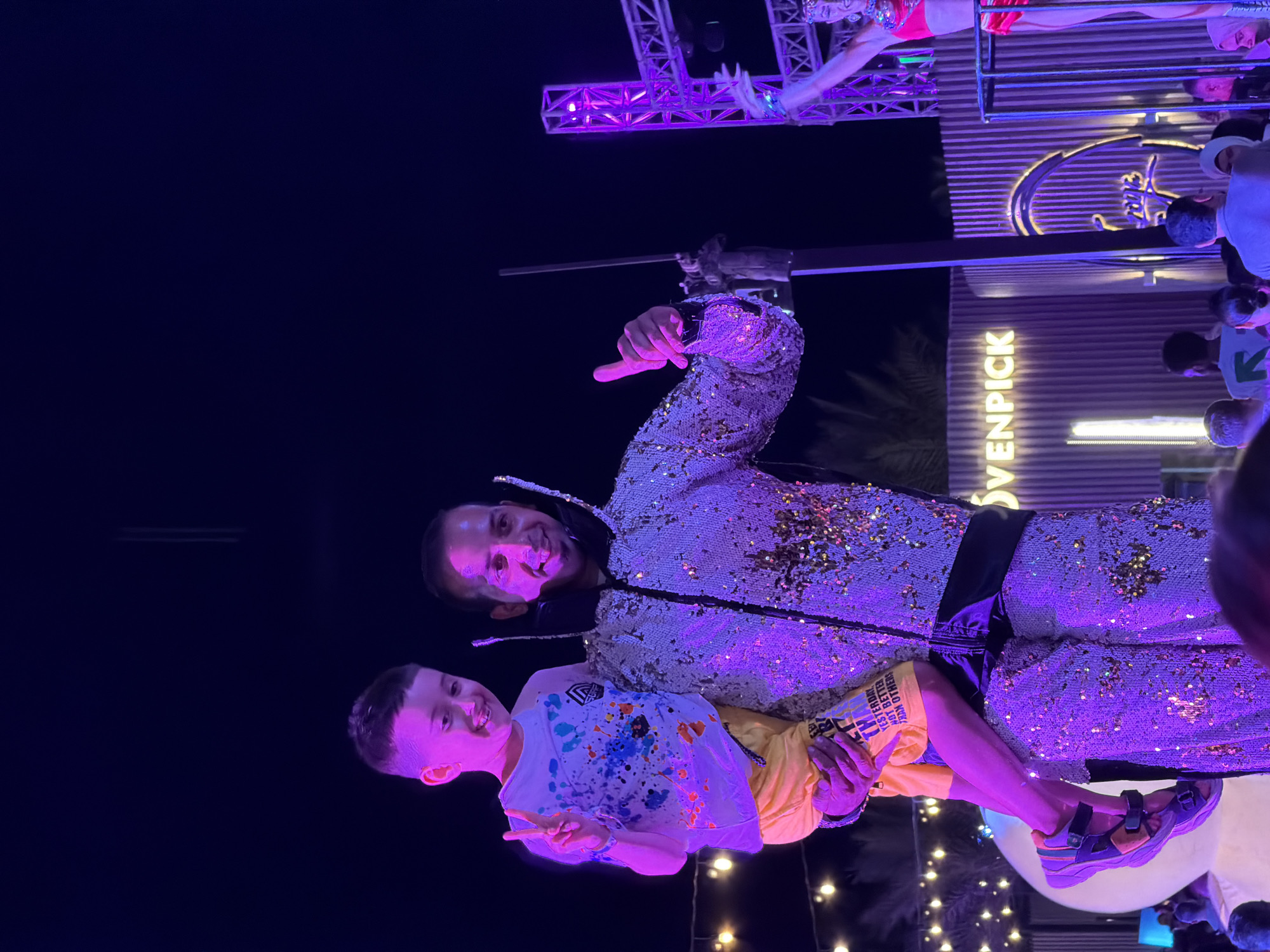 The image shows two people, an adult and a child, posing together in front of a backdrop illuminated by purple lights. The adult, wearing a sparkling sequined jacket, is holding the child, who is wearing a white t-shirt with colorful paint splatters and yellow shorts. The child is making a peace sign with their fingers and smiling at the camera. The setting appears to be an outdoor evening event, possibly a performance or a celebration, given the stage-like lighting and the presence of other blurred figures in the background. A large sign with illuminated text reading "VENPICK" is visible on a building in the background, suggesting the location is a venue or hotel. The overall mood is joyful and celebratory, with the bright lights and smiling faces. The time of day is clearly night, indicated by the dark sky and artificial lighting. The location is identified as Sharm El Sheikh, Egypt.