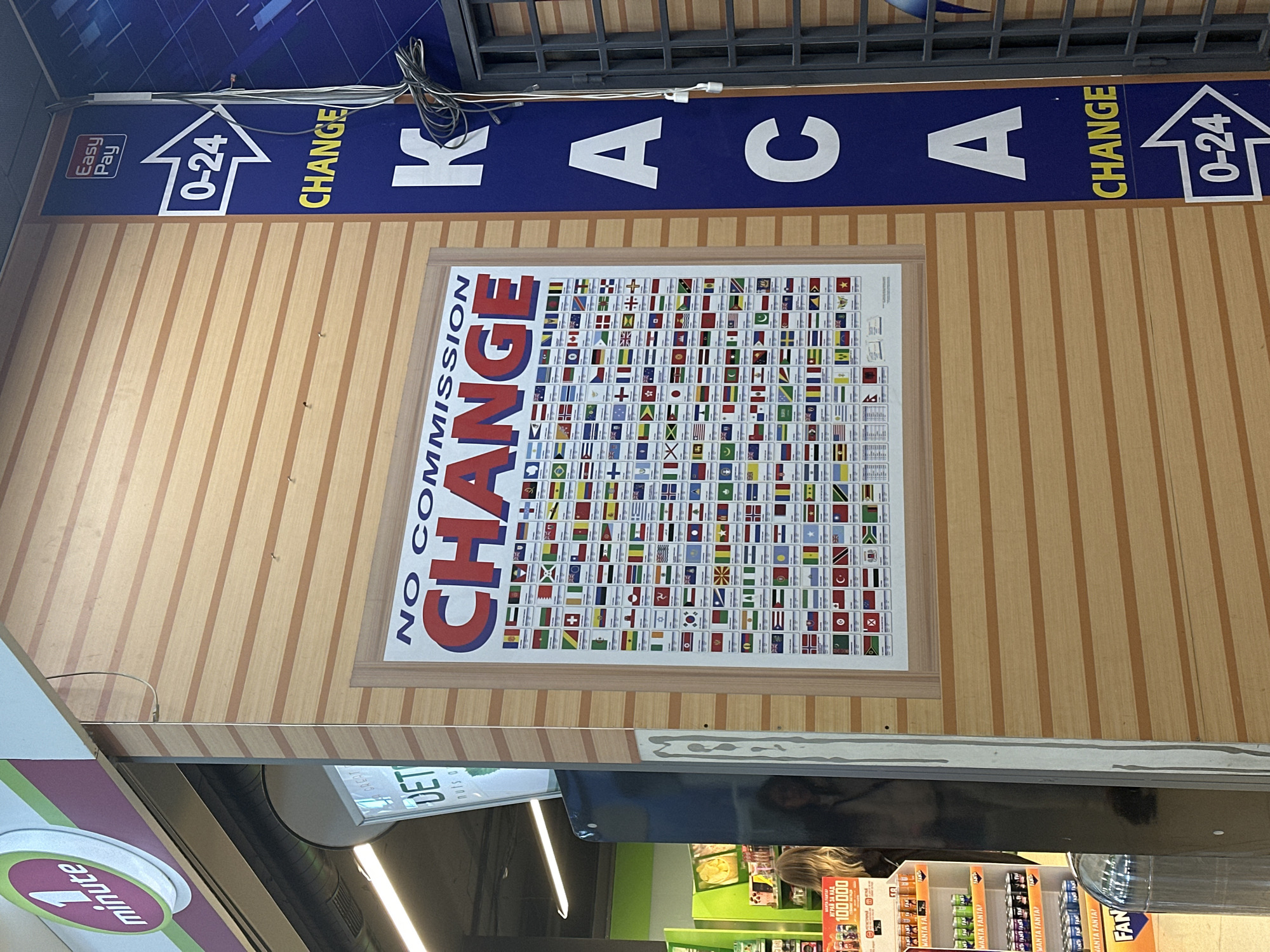 The image shows an indoor exchange office or kiosk, possibly in Sofia, Bulgaria, as implied by the Bulgarian currency exchange poster displayed. The foreground features a '1 minute' branded sign. Above, on a wall with vertical wooden paneling, is a framed poster titled "NO COMMISSION CHANGE" with a display of national flags. Above the poster, there is a blue sign that says 'KACA CHANGE 0-24 Easy Pay'. Below, the foreground shows some shelves of Fanta drinks, some unidentified printed material saying 'ИГРАЙ ЗА НАД 100000', and what appears to be a person's head reflected in a reflective surface.