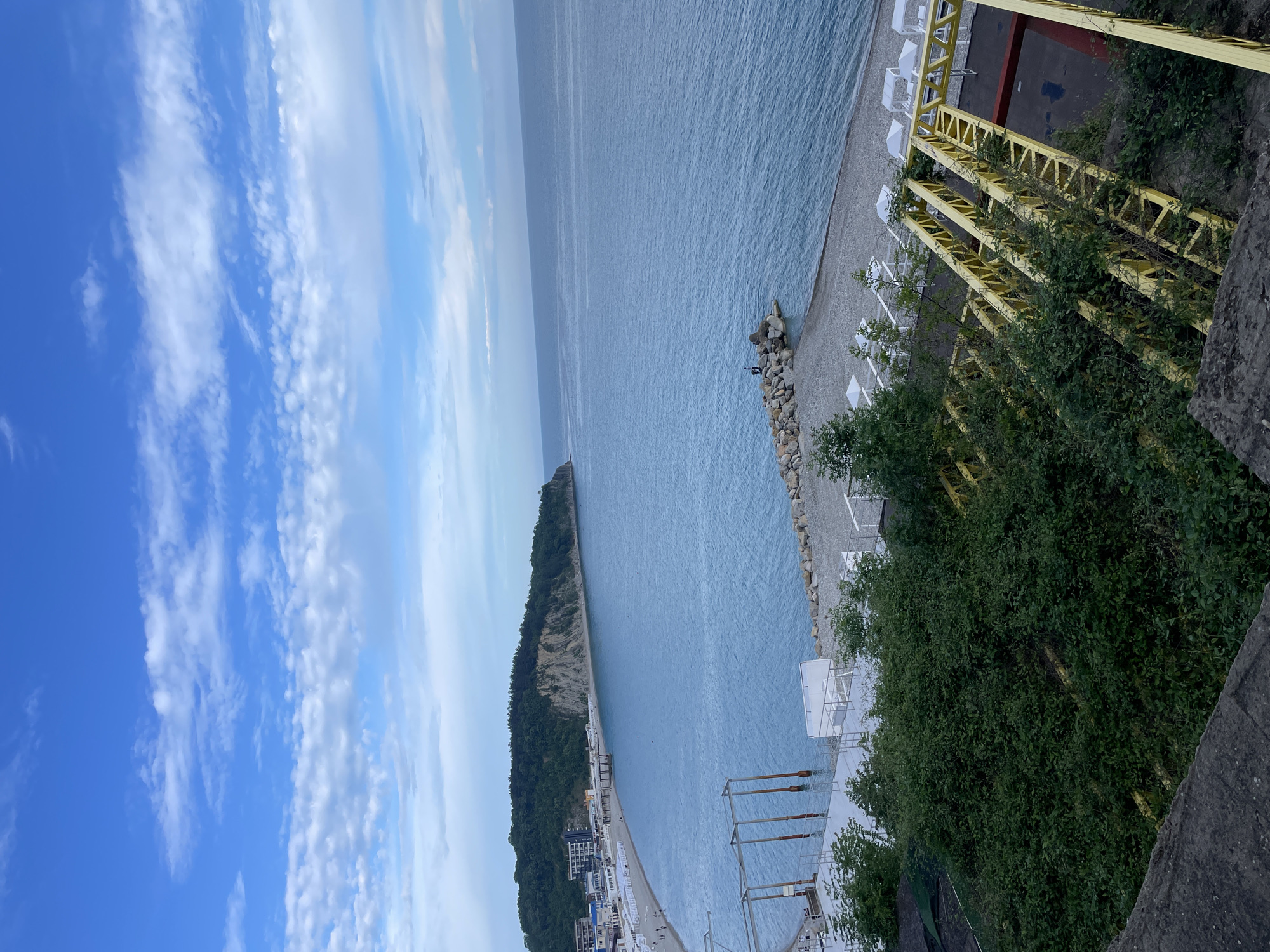 The image captures a view of the coastline near Olginka, Russia. The sea stretches out in shades of blue and grey, meeting a pebble beach lined with white sun loungers. A promontory covered in lush green forest extends into the water. A structure of yellow beams and girders is visible along the shoreline, partially obscured by foliage. In the distance, buildings can be seen along the shoreline. The sky is blue with streaks of white clouds.