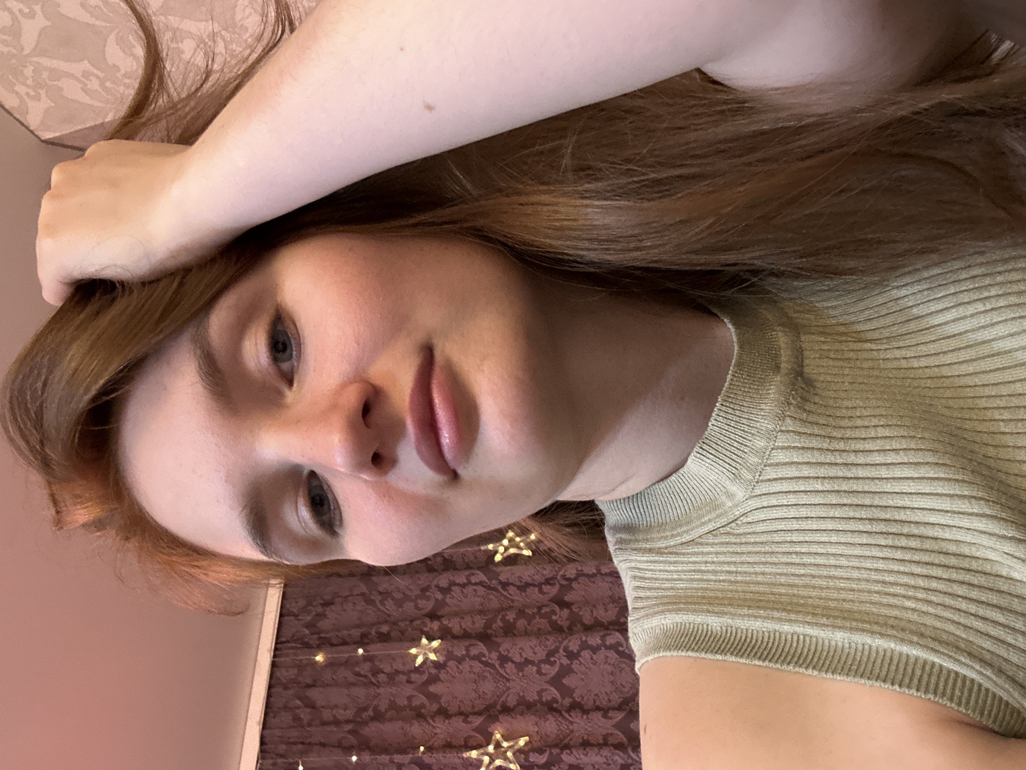 A close-up, tilted selfie of a fair-skinned young woman with long, reddish-brown hair. She is wearing a light olive green ribbed knit top. Her left arm is raised above her head, hand resting on her hair. A wall covered in dark burgundy patterned fabric adorned with small, star-shaped string lights is visible in the background. The photograph’s location in the city of Chaikovsky, Russia is not determinable from the image.