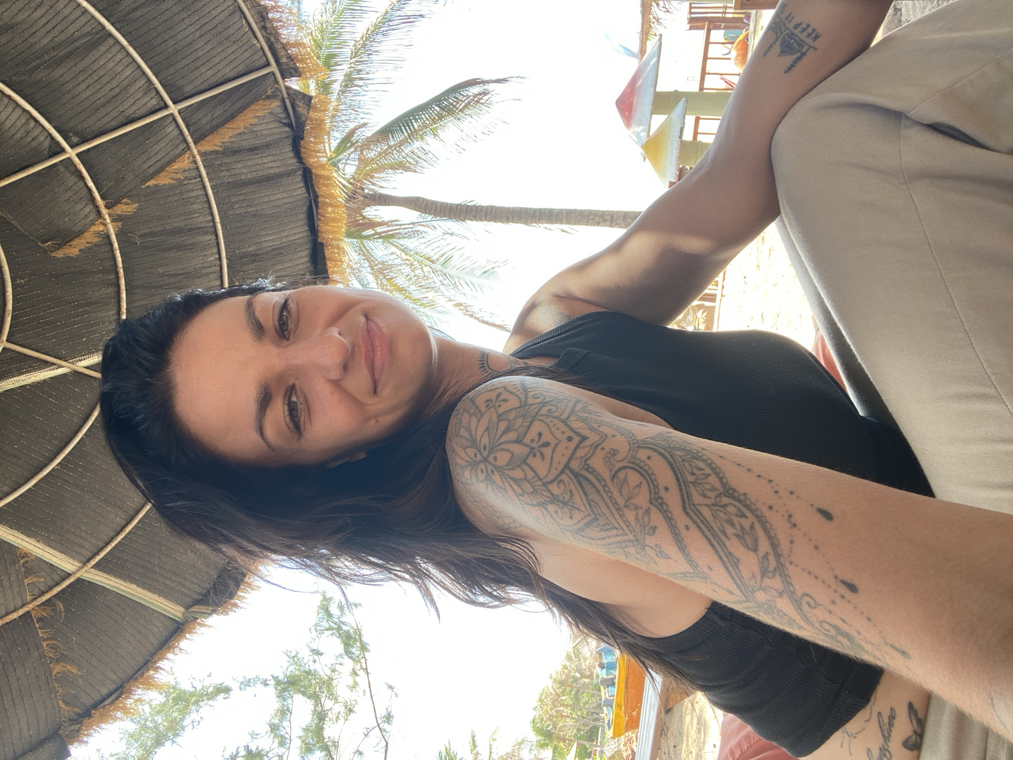 A medium-shot, eye-level, outdoor photograph shows a woman with long, dark hair and tattoos on her left arm, smiling while looking towards the camera. She is wearing a black tank top and light-colored pants. The woman is seated outdoors, under a thatched umbrella, with palm trees and a bright, overcast sky visible in the background. In the middle ground, a yellow boat rests on a wooden structure near a beach. In the far background, there are several colorful structures that appear to be part of a resort or recreational area. The overall scene suggests a relaxed, tropical environment, likely a beach resort in Mũi Né, Vietnam, given the presence of palm trees and the architectural style of the background structures.