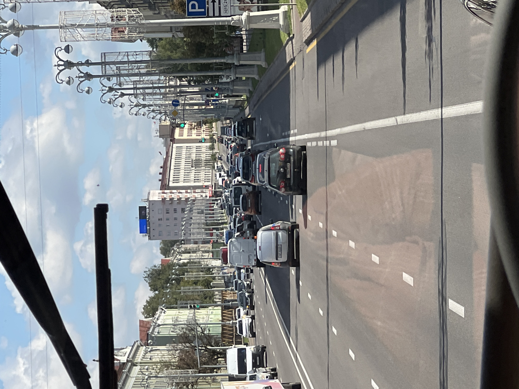 This is a photograph taken from the front of a vehicle, likely a bus or tram, looking forward along a multi-lane city street in Minsk, Belarus. The sky is blue with some white clouds. On either side of the street are buildings, some appearing to be residential or office blocks with ornate facades and arched windows, while others are more modern. Trees line the sidewalks.
The street is busy with traffic. Several cars, vans, and trucks are visible, driving in multiple lanes. There are also traffic lights and streetlights. The road surface shows lane markings and some reddish stains, possibly from recent rain or spills. The perspective suggests the vehicle is in motion, contributing to a sense of urban activity.
The composition is dominated by the street and the vehicles on it, with the cityscape forming the backdrop. The top of the image is partially obscured by dark, horizontal bars, which are likely part of the vehicle the photo was taken from, possibly overhead wires or window frames.