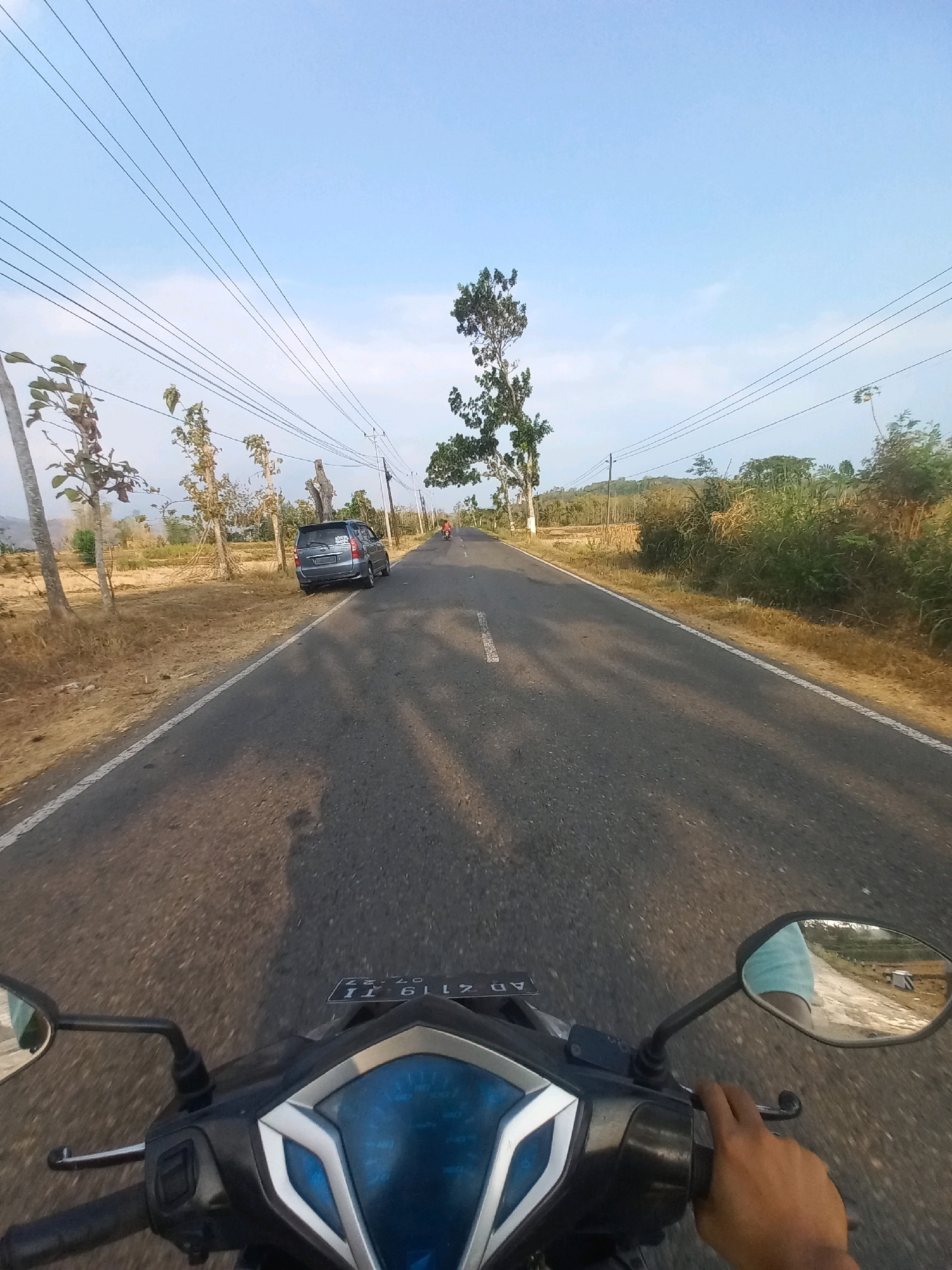 A point-of-view shot from a motorcycle traveling down a paved road in Nguntoronadi, Indonesia. The sky is clear and blue with some faint clouds. Several power lines run parallel to the road. On the left side of the road, there is dry, brown vegetation, scattered trees, and a grey car parked on the shoulder. On the right side, there is more dense, green vegetation and trees. In the distance, a lone figure on a motorcycle is visible on the road. The handlebars and instrument panel of the motorcycle the photographer is riding are in the foreground. The photographer's right hand is visible on the handlebar, wearing a light blue sleeve. The license plate of the motorcycle reads "AD 7119 TI".