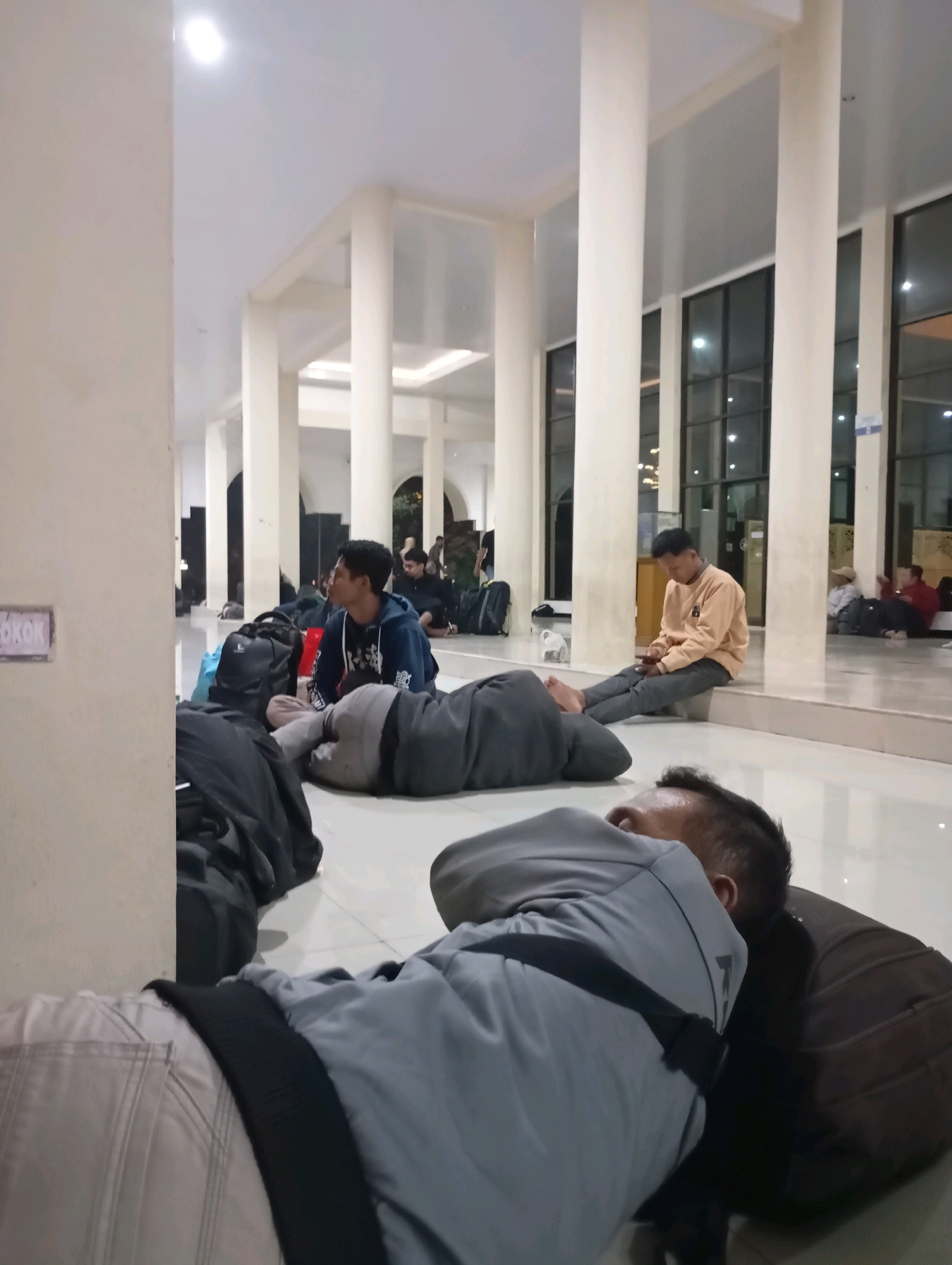 In an indoor public space, possibly a waiting area or a transit hub in Segoro Tambak, Indonesia, several individuals are resting on the floor amidst luggage. The environment features a smooth, light-colored tiled floor, large pillars supporting the ceiling, and a wall with windows.
In the foreground, two men are lying down. The man closest to the camera is on his side, facing away, wearing a grey long-sleeved shirt and light-colored pants. A dark strap crosses his chest. Next to him, a second man is also lying down, wearing a grey top and dark pants.
Further back, a man in a blue hoodie is seated, looking to his left, with several bags placed around him. To his right, another man is seated on a raised ledge, wearing a light orange long-sleeved shirt and dark pants, looking down at his phone.
In the background, more people are seated or lying down, and various bags are scattered throughout the space. The lighting appears to be artificial, originating from ceiling fixtures. A sign with the word "ROKOK" (tobacco in Indonesian) is visible on a wall to the left. The overall scene suggests a moment of repose for travelers or individuals waiting for an extended period.