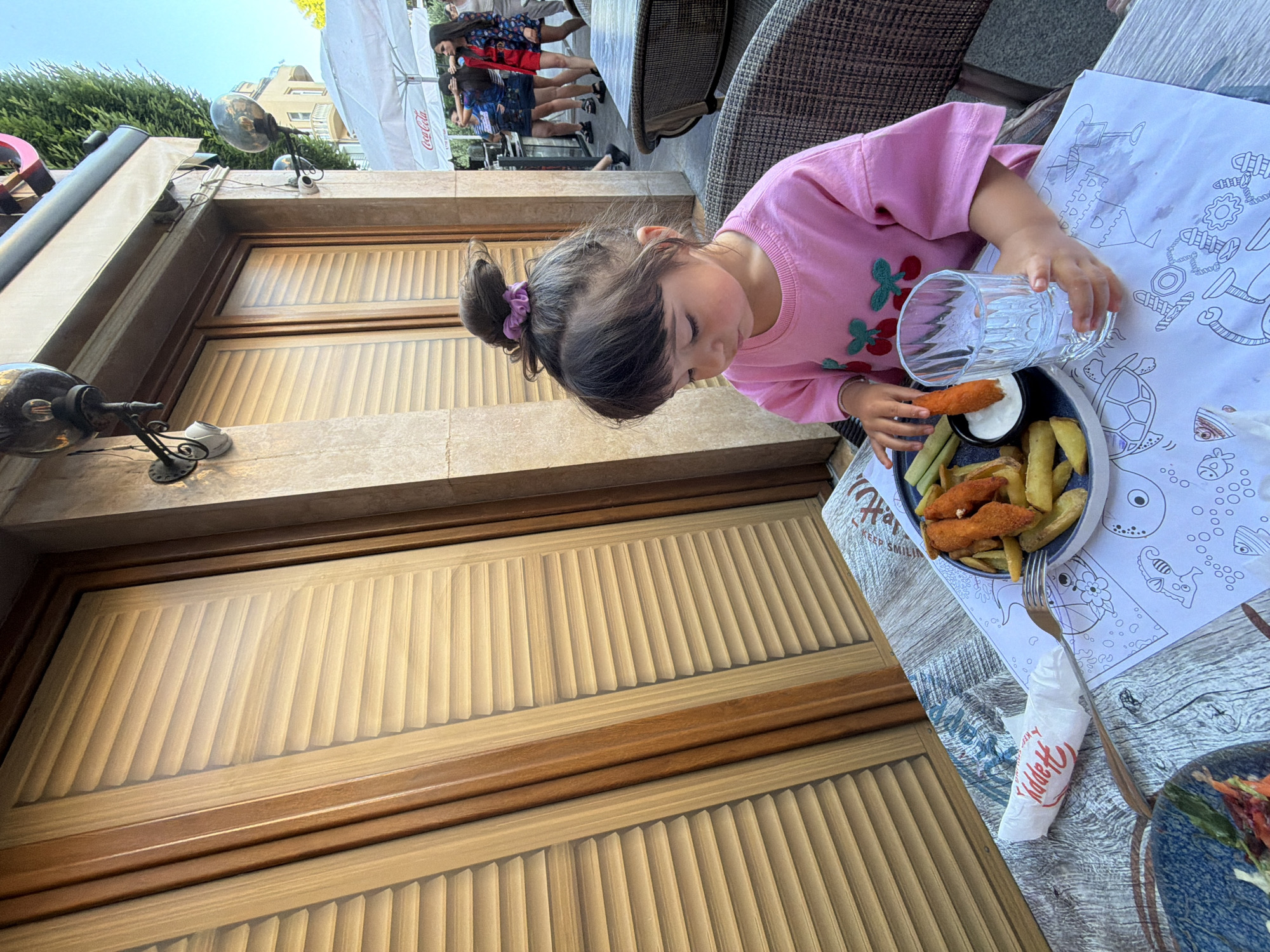 A young girl with dark hair pulled into a bun, wearing a pink t-shirt with a cherry design, sits at a table outdoors at a restaurant in Burgas, Bulgaria. She is holding a glass of water and reaching for a piece of food, which appears to be a chicken tender, from a plate of chicken tenders and french fries. A napkin with the word "Happy" printed on it is on the table, along with a fork. The table has a printed paper placemat with drawings of sea creatures and a castle. In the background, there are wooden shutters on a building, outdoor seating, a white umbrella with "Coca-Cola" printed on it, and several adults walking by. The lighting suggests it is daytime.