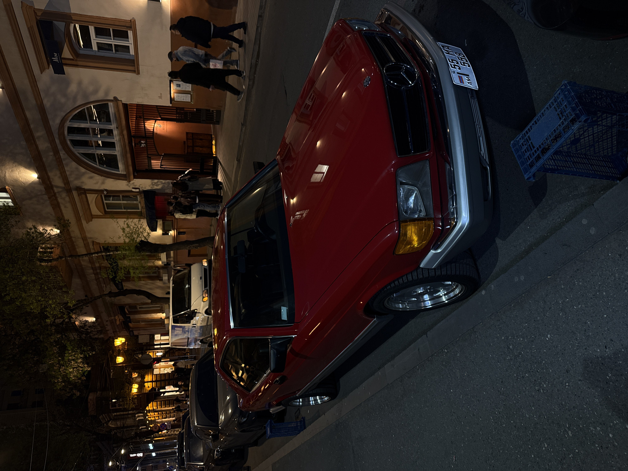 The image shows a street scene at night in Yerevan, Armenia. A red vintage Mercedes-Benz is parked along the right side of the street. A blue plastic crate sits near the car's front. Several people are walking along the sidewalk on the left side of the street in front of a building with architectural details and lit windows. Streetlights are visible in the distance, as are other parked vehicles. The Armenian license plate 55 MA 555 is visible on the vehicle.