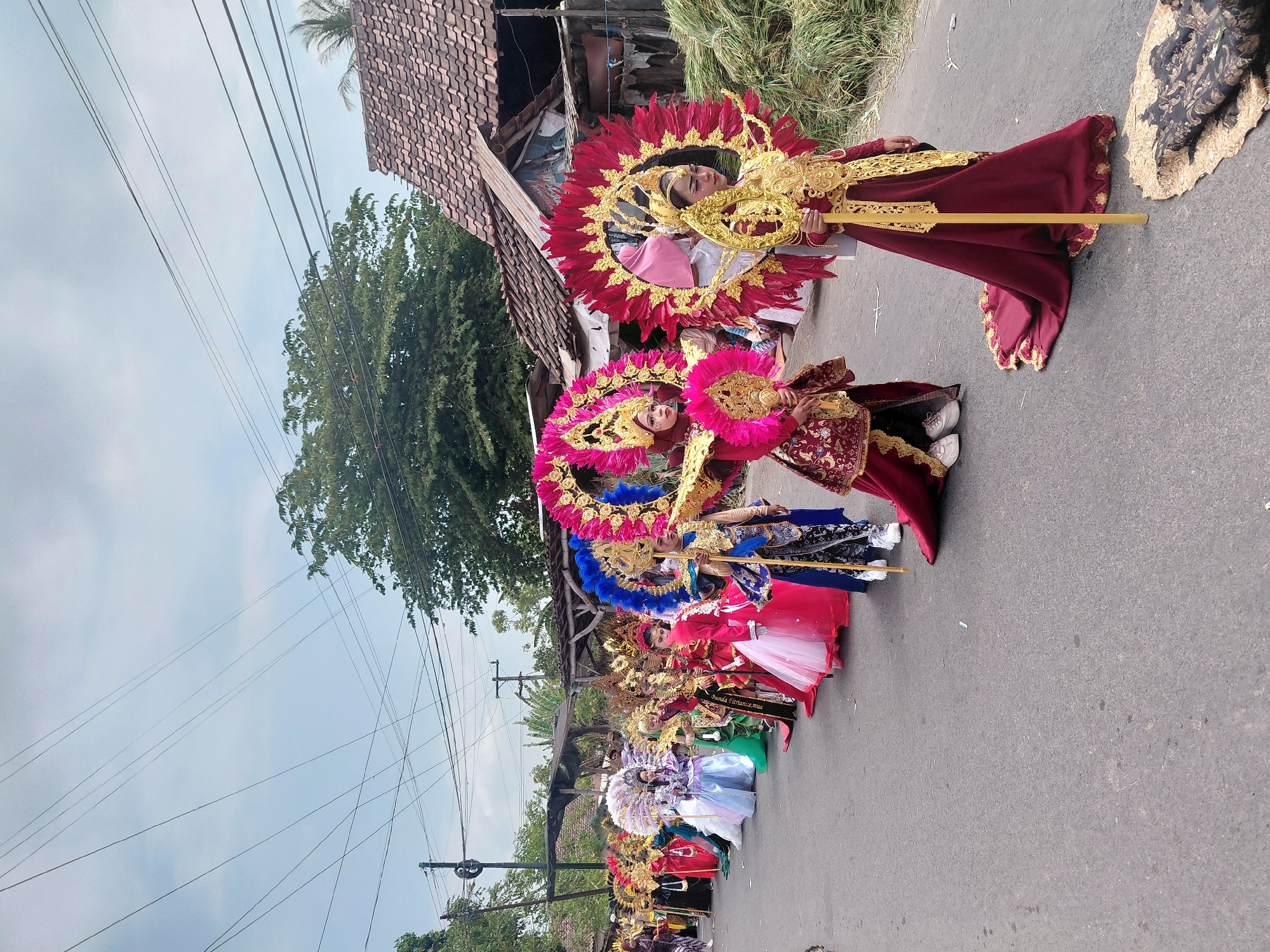 In Bimo, Indonesia, a procession of young people in elaborate costumes walks down a paved street. The participants, mostly female and wearing vibrant, ornate attire, are adorned with large, colorful headdresses featuring feathers and gold accents. Some carry staffs or fans. Their costumes include richly colored fabrics like red, blue, pink, and white, with intricate gold embroidery and embellishments. The procession is moving from left to right in the frame, with the sky overhead filled with scattered clouds and a dense network of overhead power lines. On the right side of the street, there are houses with tiled roofs and piles of what appears to be harvested vegetation or hay. The overall scene depicts a cultural event or parade, likely a celebration or festival.