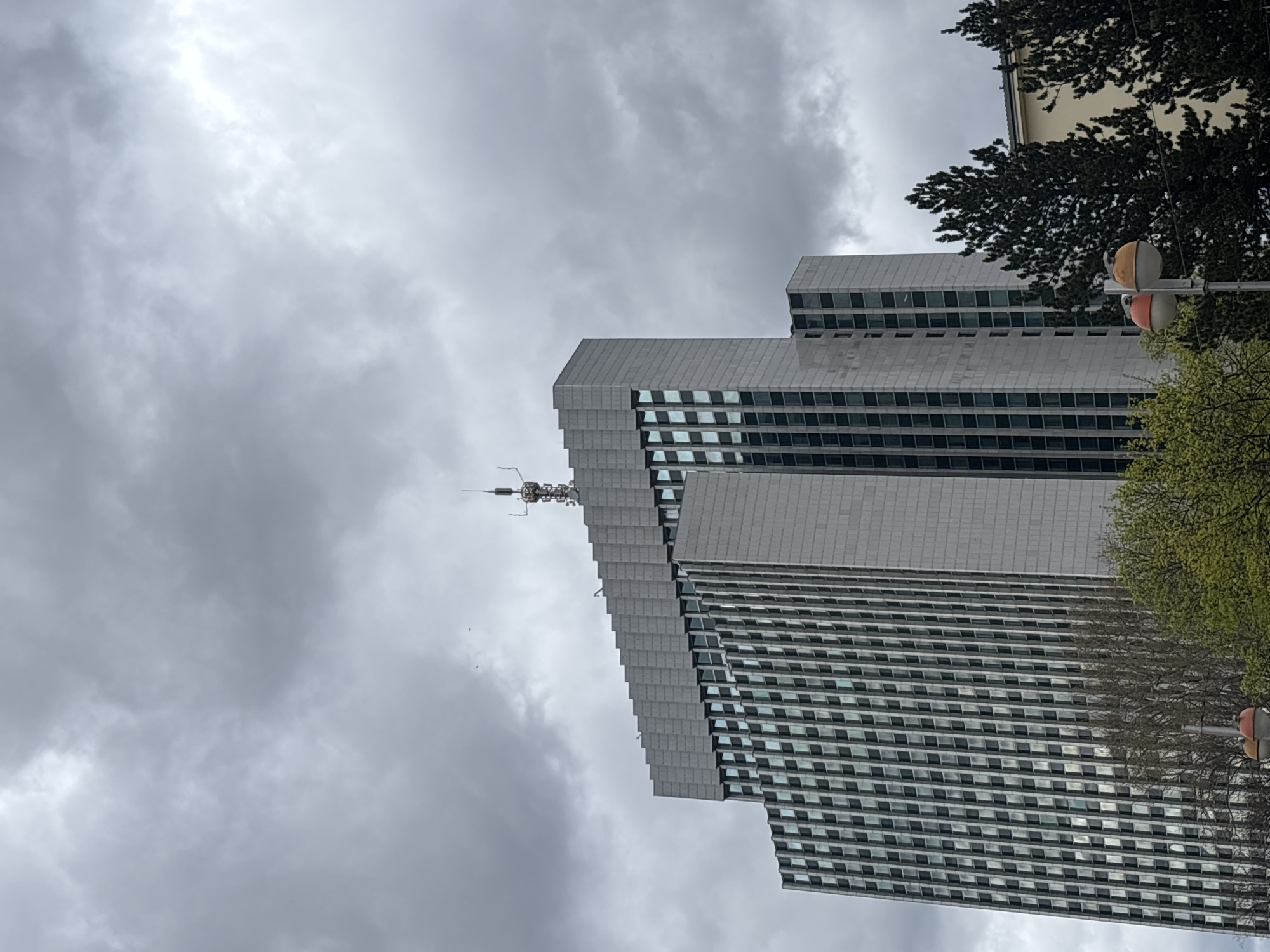 The image shows the Zagrebtower in Zagreb, Croatia. The building is tall with a distinctive stepped design and a broadcast antenna on top. The sky is overcast with gray clouds. Trees are visible to the right of the tower. A traffic light is also seen near the trees.