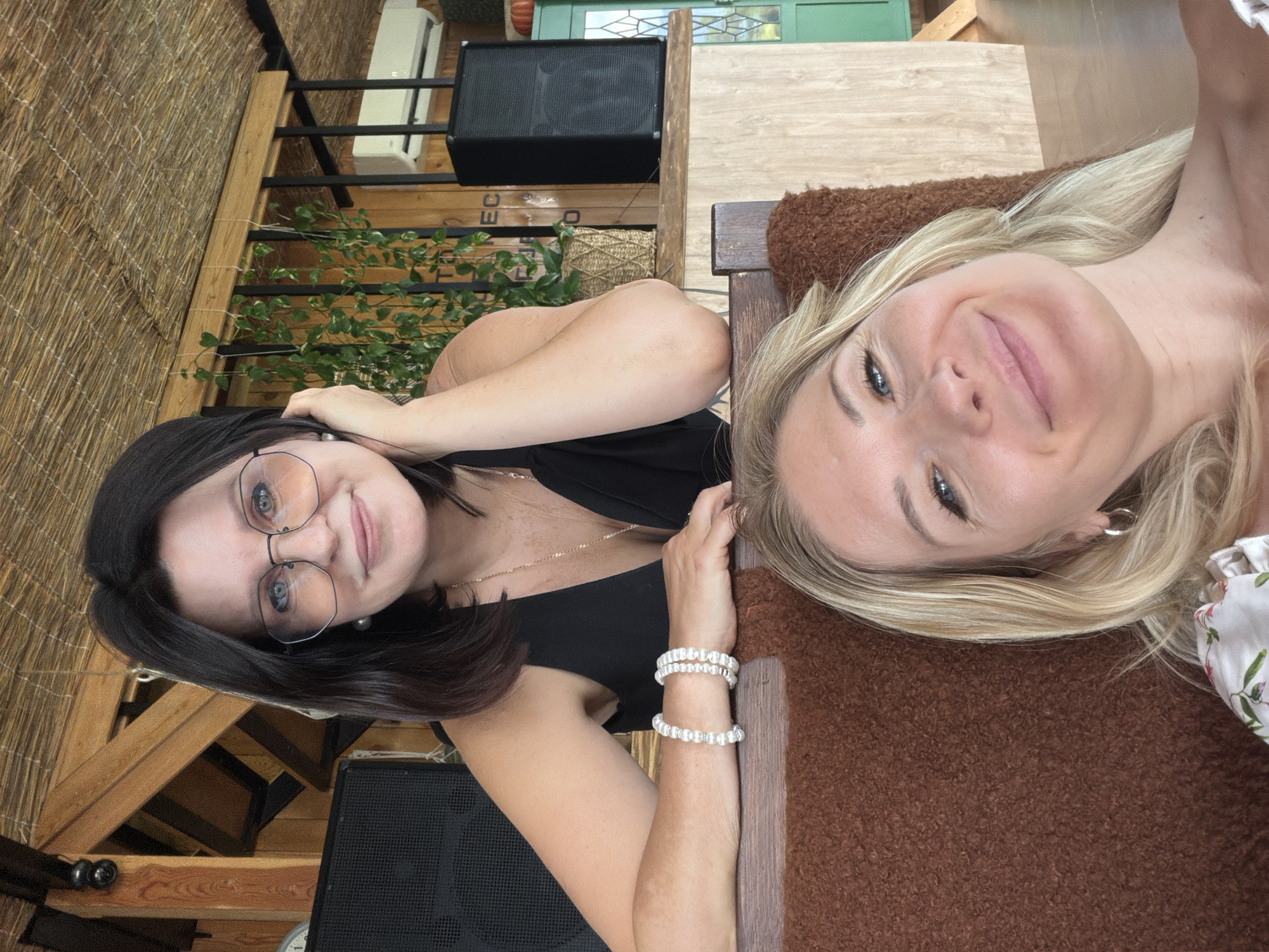 In Nizhnyaya Shilovka, Russia, two women are pictured. The woman in the foreground has blonde hair and is smiling at the camera. She is wearing a floral-patterned top. Behind her, a woman with dark hair and glasses is leaning on her hand and also looking towards the camera. She is wearing a black dress and several bracelets on her arm. The background suggests an indoor setting with wooden beams, a thatched ceiling, plants, speakers, and an air conditioner unit.