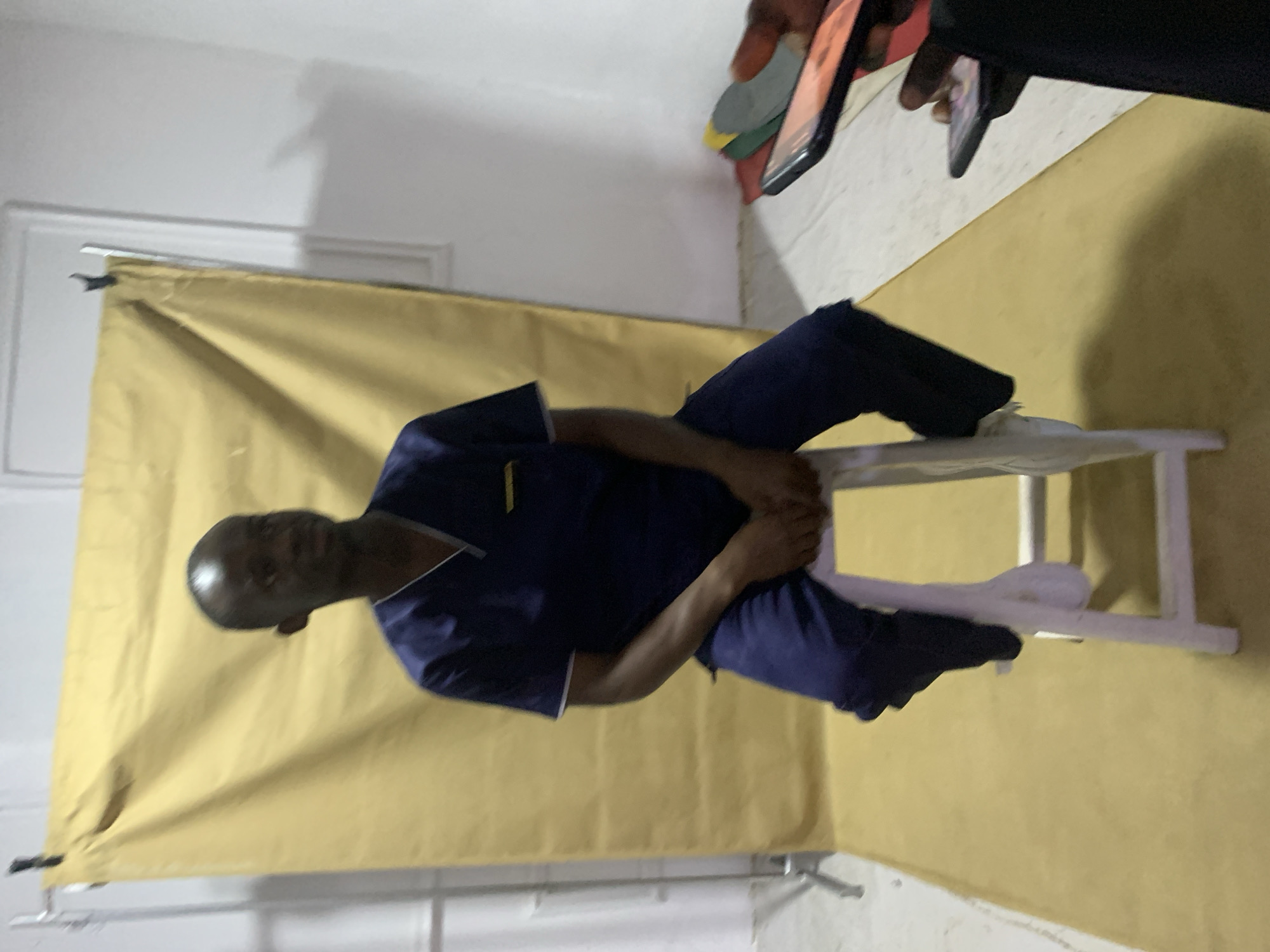 The image shows a man sitting on a white stool in front of a yellow backdrop. He is wearing a dark blue scrub suit with white trim. He is facing left and has his hands clasped in his lap. The backdrop is wrinkled. Two people with phones are visible in the upper right corner of the image. The location is stated to be in Jos, Nigeria. The environment appears to be indoors, likely a studio or makeshift photo area.