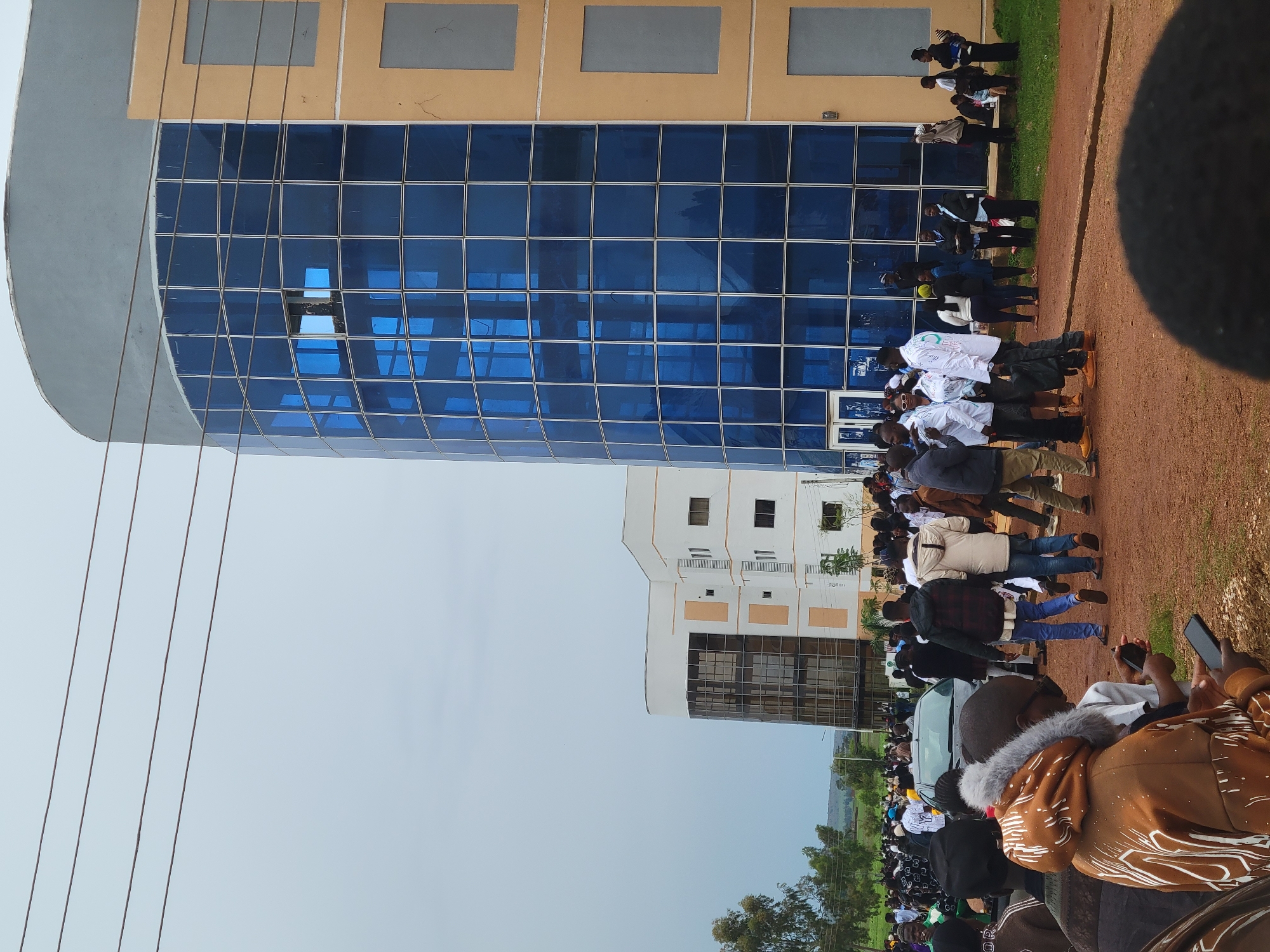 The image depicts an outdoor scene with a large group of people gathered in front of modern buildings, one of which features a striking blue glass facade and a curved upper section. The ground is a mix of reddish-brown dirt and patches of green grass. In the background, a second, white building with a similar architectural style is visible. The sky is overcast.
The people are dressed in a variety of casual and semi-formal attire. Some are standing and conversing in small groups, while others are walking or looking at their phones. The composition suggests a lively gathering, possibly a campus event or public assembly. There are vehicles present, including a car in the middle ground. Electrical wires are visible in the upper part of the frame.
The location is identified as Durumi, Nigeria. The architecture of the buildings, characterized by large glass panels and contemporary design, points to a developed urban or institutional setting within the city. The presence of numerous people indicates a degree of social activity or organized event.