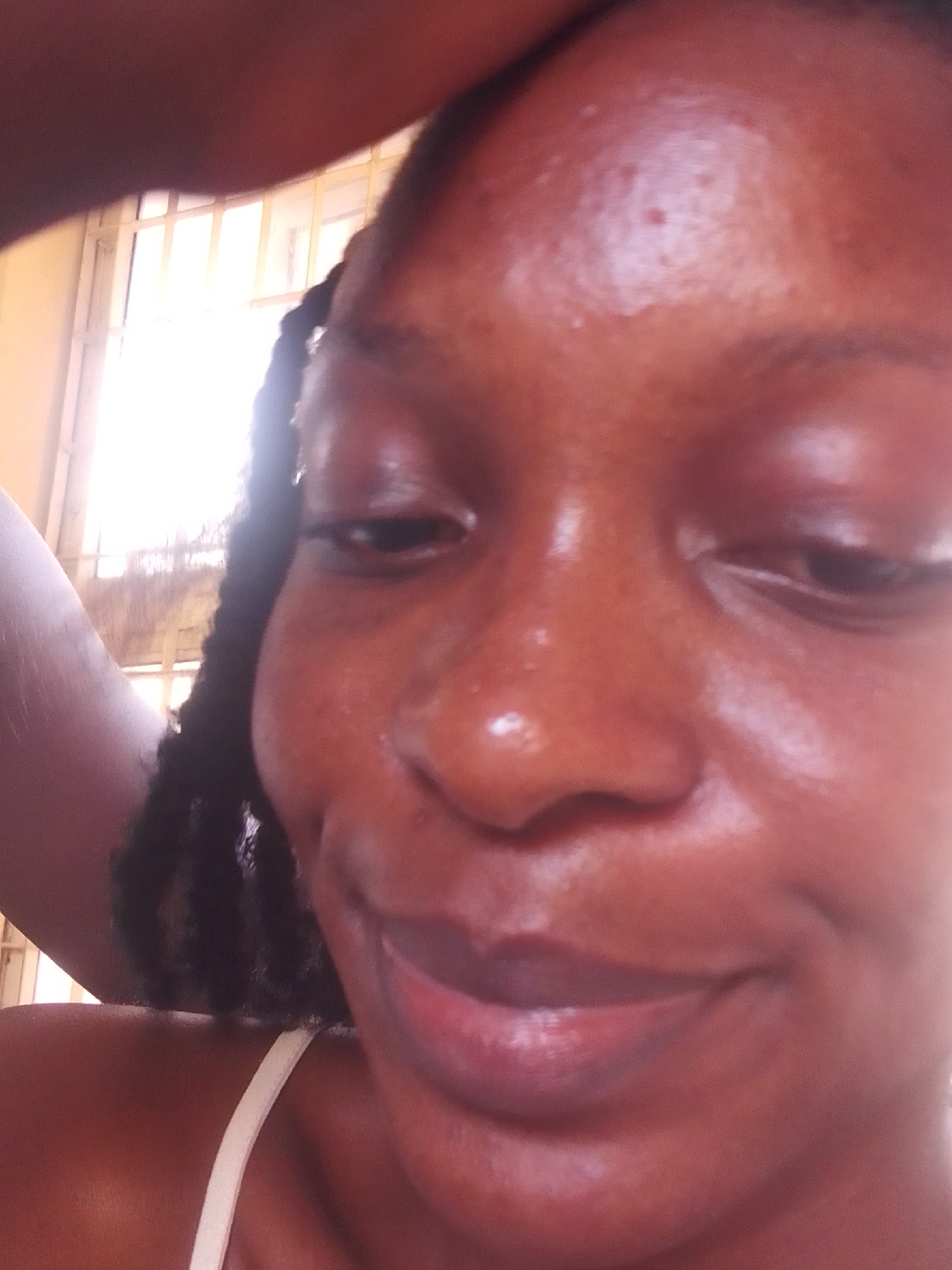 The image is a close-up, frontal selfie of a young Black woman with braided hair. She is looking slightly down and to her right, with a subtle smile. Her skin appears to be glowing, possibly due to natural oils or a skincare product. There are some small blemishes on her forehead. She is wearing a white strap, likely from a tank top or bra. In the background, out of focus, is a window with vertical bars. The lighting is bright, suggesting daylight. The overall impression is a candid, unposed moment captured by the subject herself.
