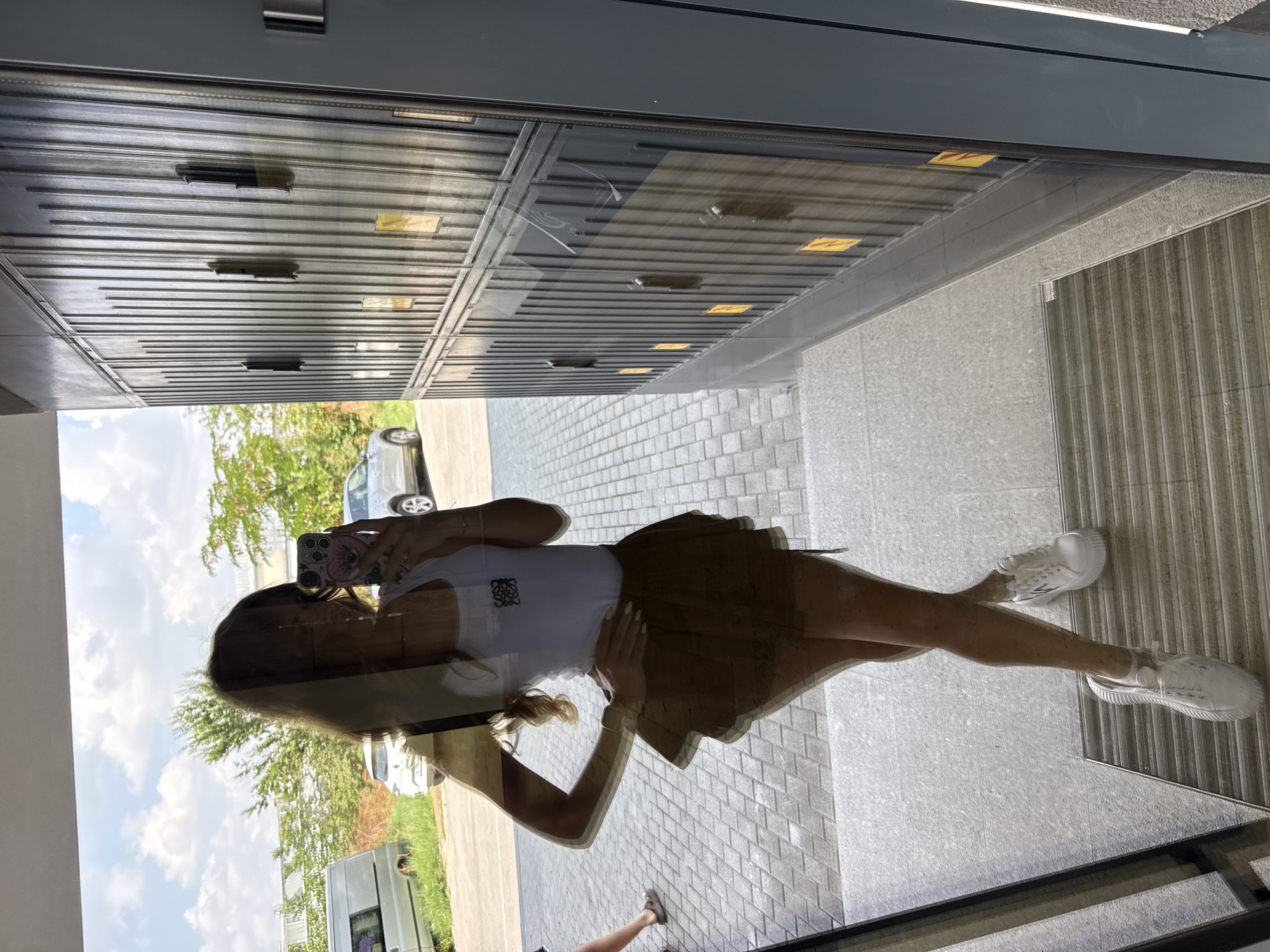 A young woman with long, light brown hair is reflected in a glass surface. She is wearing a white tank top with a black logo, a brown tiered mini-skirt, and white sneakers. She is holding a pink smartphone in her right hand, which is reflecting in the glass. Her left hand is on her hip. She is standing on a paved walkway in front of a building with a row of dark, ribbed lockers. Outside, there are trees and parked cars, including a silver SUV and a white van, visible under a cloudy sky. This scene is located in the city of Plovdiv, Bulgaria.