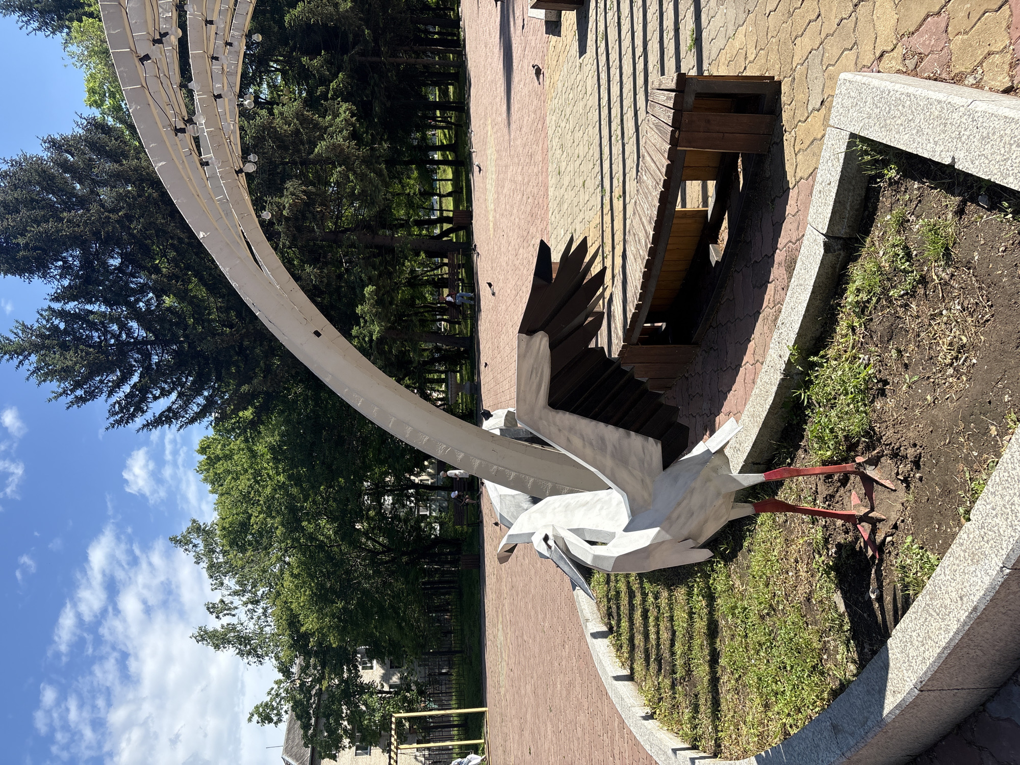 In Birobidzhan, Russia, a modern public park features a striking sculpture of a stork. The stork, rendered in a stylized, abstract manner with sharp angles and a metallic sheen, is positioned in a bed of dirt and grass bordered by granite-like curbing. Its white body contrasts with its red legs and dark wingtips. Behind the sculpture, a wooden bench is partially visible, casting a shadow on the patterned paving stones of the park. A white, arched canopy structure, strung with lightbulbs, extends overhead, suggesting it's an area for public gatherings or events, particularly at night. Lush green trees form a dense backdrop, with a few scattered pigeons on the pavement. In the distance, a yellow children's swing set and a low fence are discernible near a multi-story building. The sky is a clear blue with fluffy white clouds, indicating fair weather.