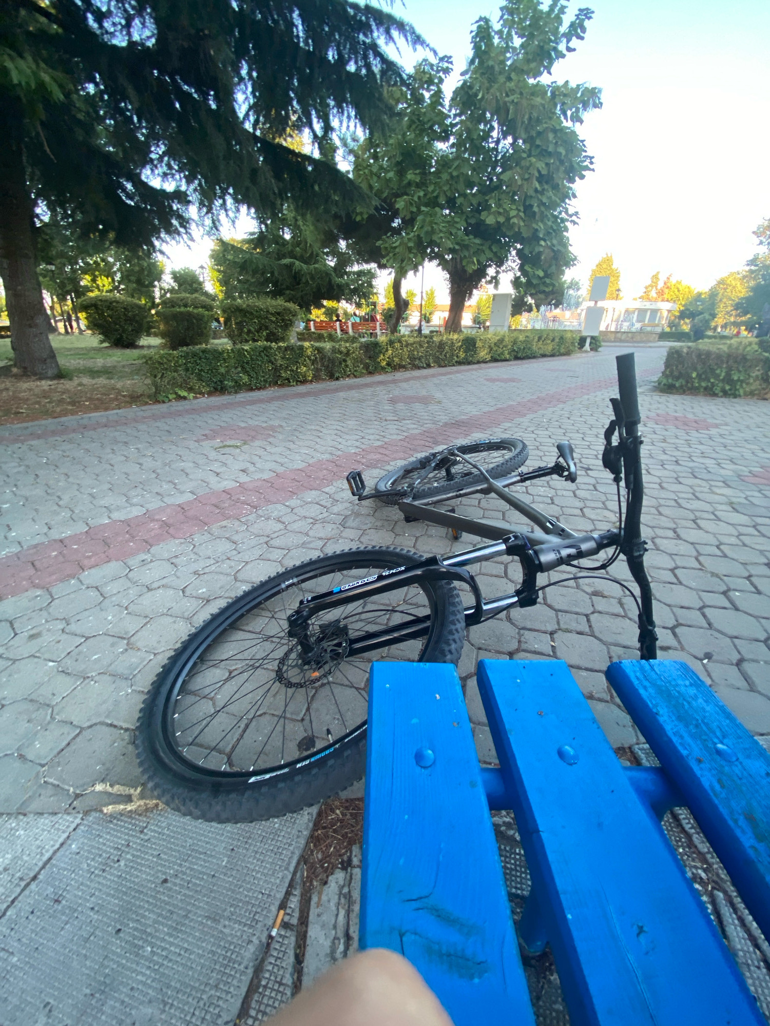 A bicycle is lying on its side on a paved walkway in a park in Pomorie, Bulgaria. The bicycle is dark grey and has disc brakes. It is positioned next to a bright blue wooden bench. The walkway is made of grey interlocking pavers with a pink stripe running through it. In the background, there are trees, hedges, and some structures, possibly part of a fountain or memorial. The sky is bright blue. The image is taken from a low angle, with a person's leg visible in the lower right corner.