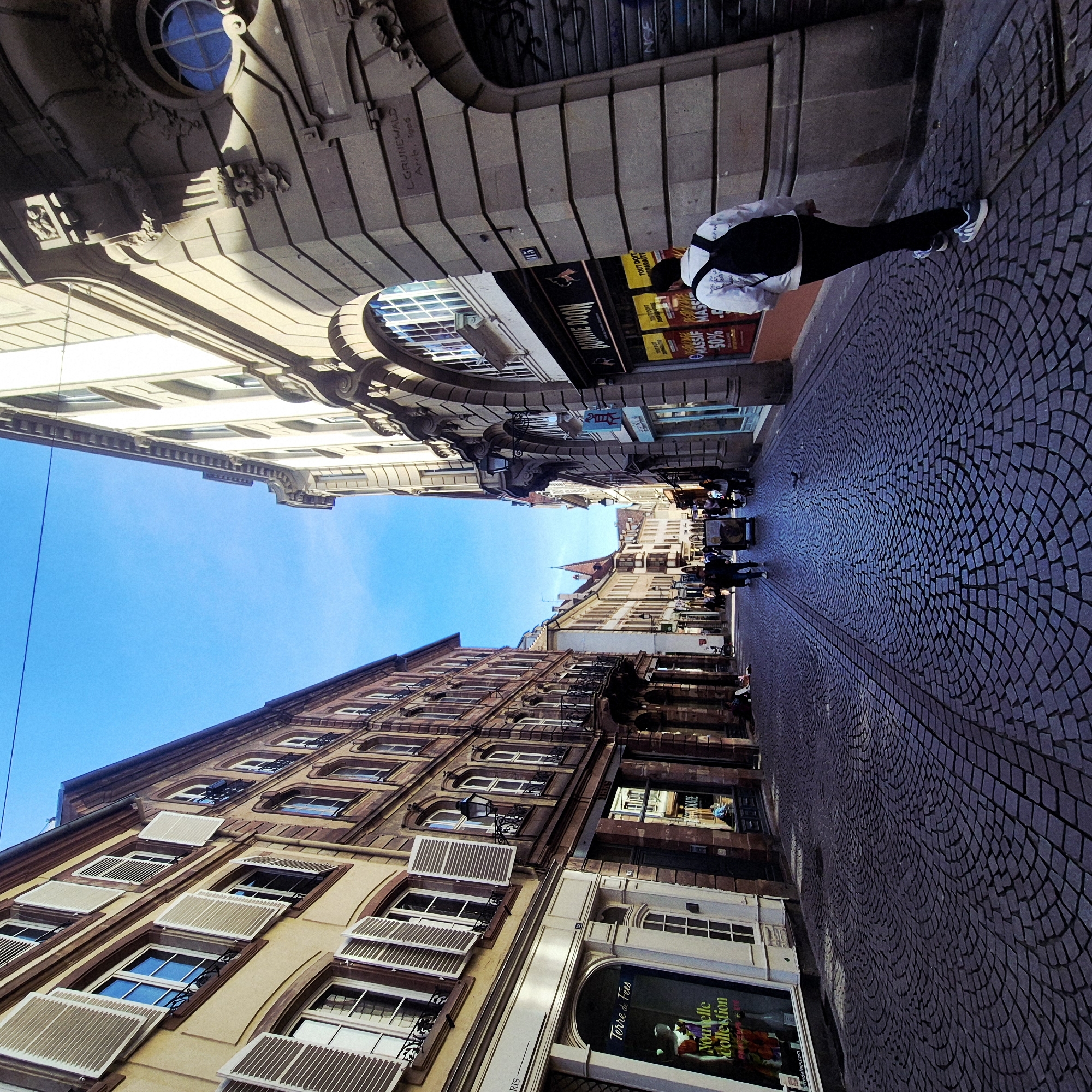 The image depicts a street scene in Strasbourg, France. The camera is positioned low and tilted, creating a perspective looking upwards along a narrow street lined with multi-story buildings. The cobblestone street is visible in the foreground, leading into the depth of the image. A person wearing dark pants and a white shirt with a black backpack is walking along the street. On the upper right, an archway spans above the street. Storefronts are on either side of the street. The sky is visible between the buildings, clear and blue. Architectural details of the buildings are apparent, including window shutters and ornamental features. Text from the storefront signs is partially legible, indicating a "Nouvelle Collection" and a "50%" offer. The building’s upper right is engraved with, “L. GRUNEWALD Arch. 1906”.