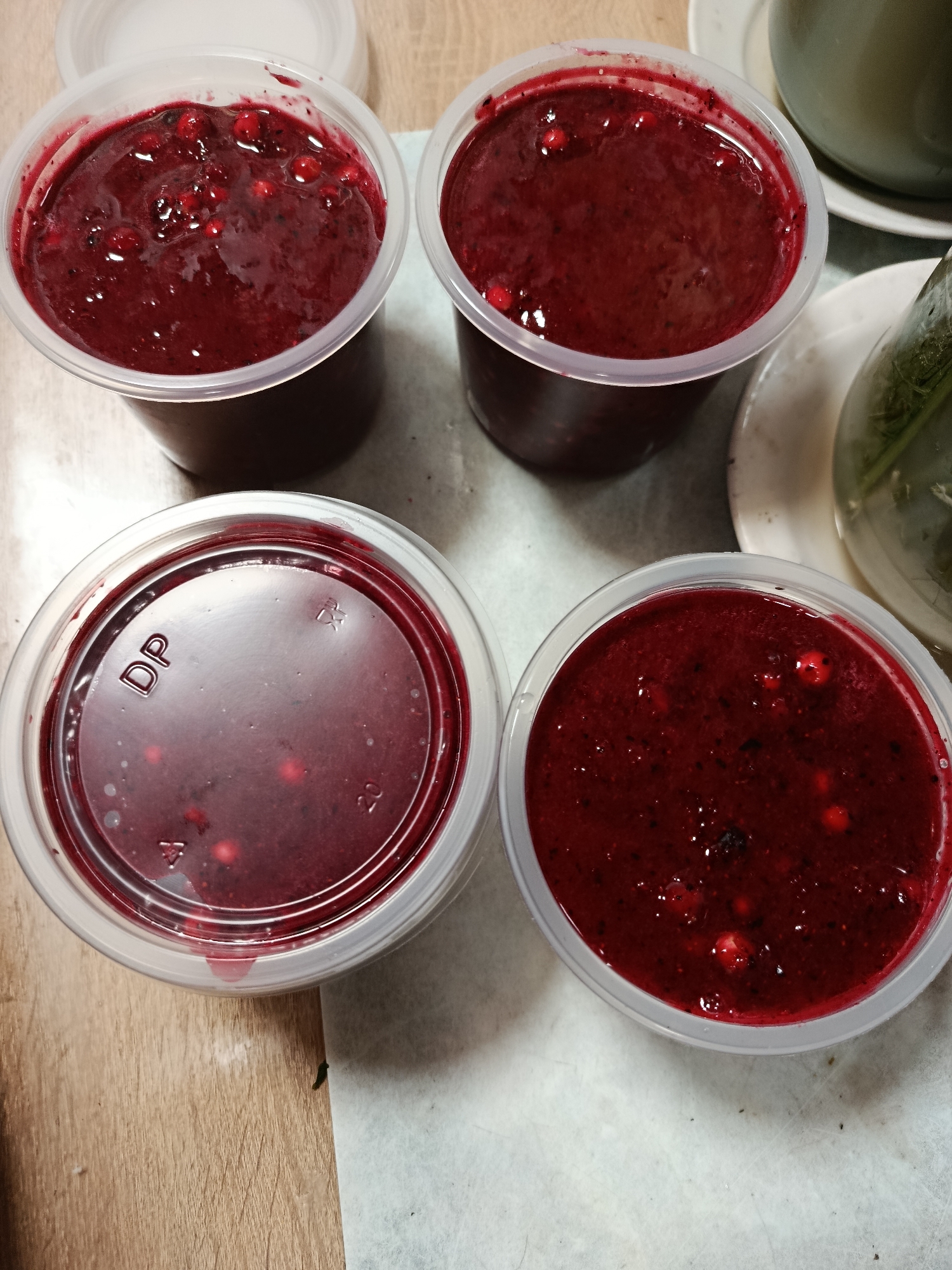 Four clear plastic containers filled with a deep red, pureed fruit substance, likely a jam or compote, are arranged on a light-colored, textured surface. The containers are round and appear to be disposable. Each container has a small amount of the red substance splattered on the inside rim. One container in the bottom left has the letters "DP" printed on its lid. In the background, to the right, is a glimpse of a pale green ceramic mug and a white saucer, suggesting a possible beverage setting. The overall scene is simple and focuses on the food items. There is no information to determine the location in the city of Верея, Russia from the image itself.