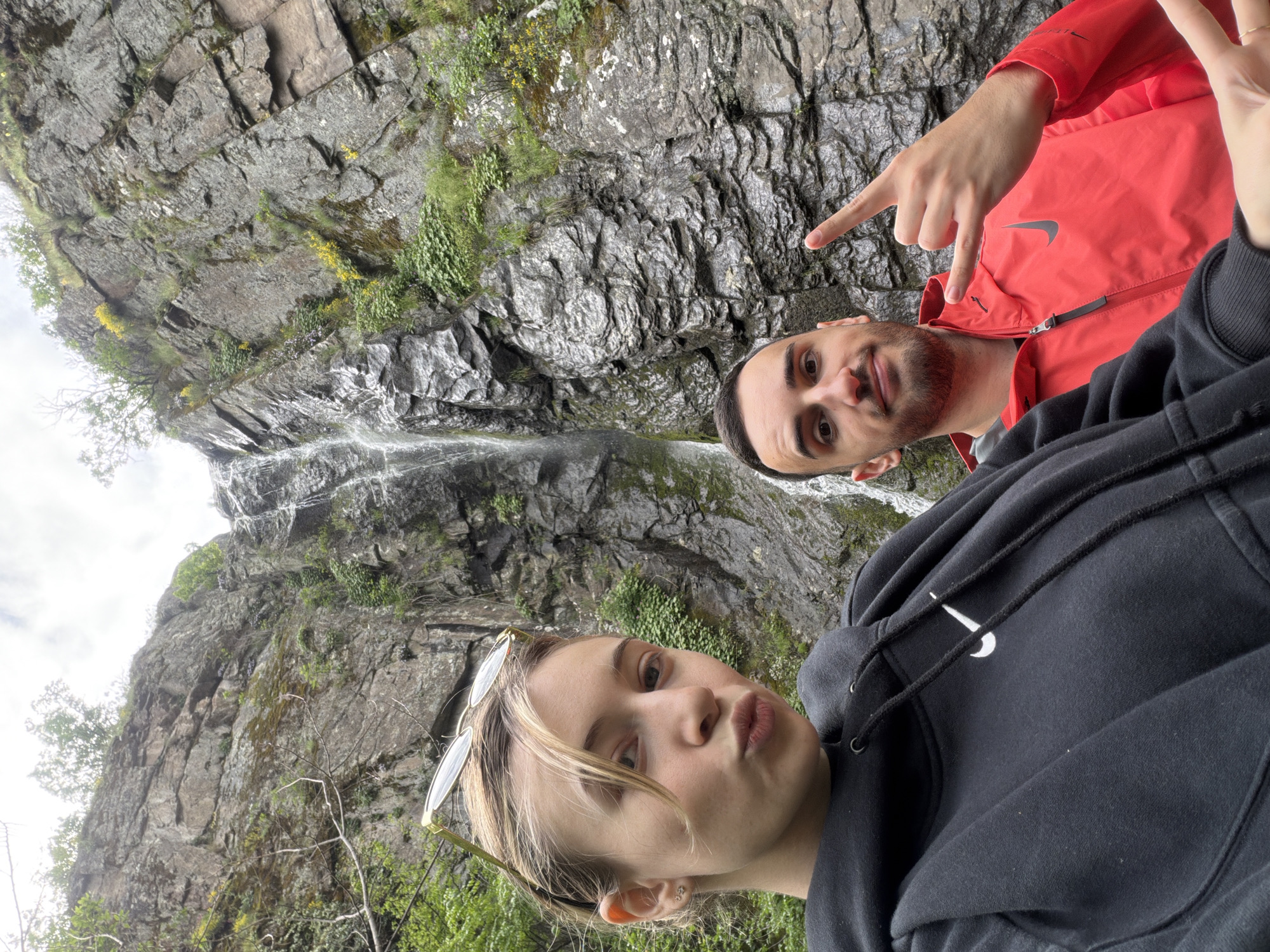Two individuals are posing for a selfie in front of a waterfall. A person wearing a black Nike hoodie is on the foreground, looking directly at the camera, while another one is on the background, wearing a red jacket and pointing to the waterfall. The background shows a waterfall over a rocky cliff with vegetation. The location is near Ustina, Bulgaria.