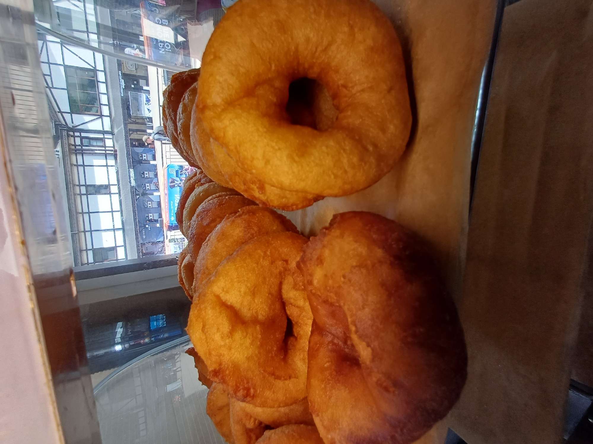 Several golden-brown, ring-shaped pastries, resembling donuts or large fried bagels, are displayed on a light brown surface, likely parchment paper or a mat, within a glass case. The pastries vary slightly in color, with some appearing a rich golden hue and others a deeper, more reddish-brown, suggesting different levels of frying or slight variations in ingredients. They are stacked in rows, creating an appetizing and abundant display.
The image is captured through the glass of the display case, which reflects an outdoor scene. This reflection shows a street or commercial area with multi-story buildings featuring numerous windows and dark frames. A person, possibly a pedestrian or customer, can be vaguely seen standing outdoors in the reflection. Various advertising signs are also visible in the reflections on the glass. Partial text from these signs includes English words like "BLUE QI CANDLEN" (likely "CANDLE IN" or similar) and "POSSESSORS" (possibly "ACCESSORIES"), as well as Cyrillic characters such as "ИВ" and "РОЗА."
The overall setting suggests a bakery, cafe, or market stall in Estosadok, Russia, where these pastries are offered for sale during the daytime, as indicated by the ambient light in the reflections. The image is oriented vertically, causing the reflected outdoor scene to appear rotated on its side.