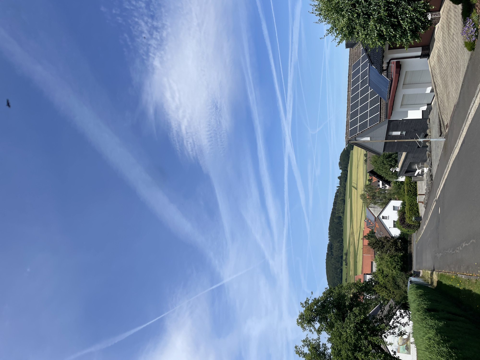 The image depicts a view of a rural settlement in Schwarzenborn, Germany, under a bright blue sky filled with numerous contrails and wispy clouds. In the foreground, a paved road curves gently to the left, bordered by a neatly trimmed green hedge on the left and a residential property with a paved driveway and a modern house featuring solar panels on its roof on the right. The house on the right has a dark gray facade and a white section with two garage doors. Beyond the immediate foreground, the scene opens up to reveal a cluster of houses with red-tiled roofs nestled among lush green trees. In the background, rolling hills covered in agricultural fields, showcasing patches of yellow and green, stretch towards a tree-covered hill under the vast sky. A small dark object, possibly a bird or insect, is visible in the upper portion of the sky.
