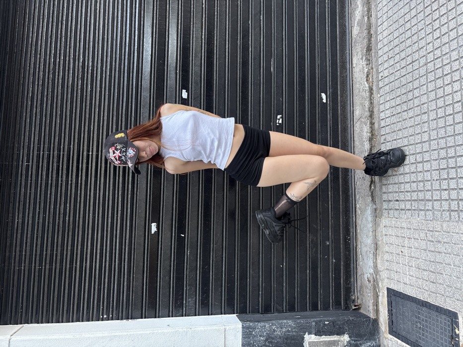 A young woman lies at an angle on a black metal grate. She wears a white tank top, black shorts, a baseball cap with designs, black sheer socks, and chunky black sneakers. Part of her body rests on the sidewalk, which is made of gray tiles with a square access point. A white curb is visible on the left. The woman looks toward the camera.