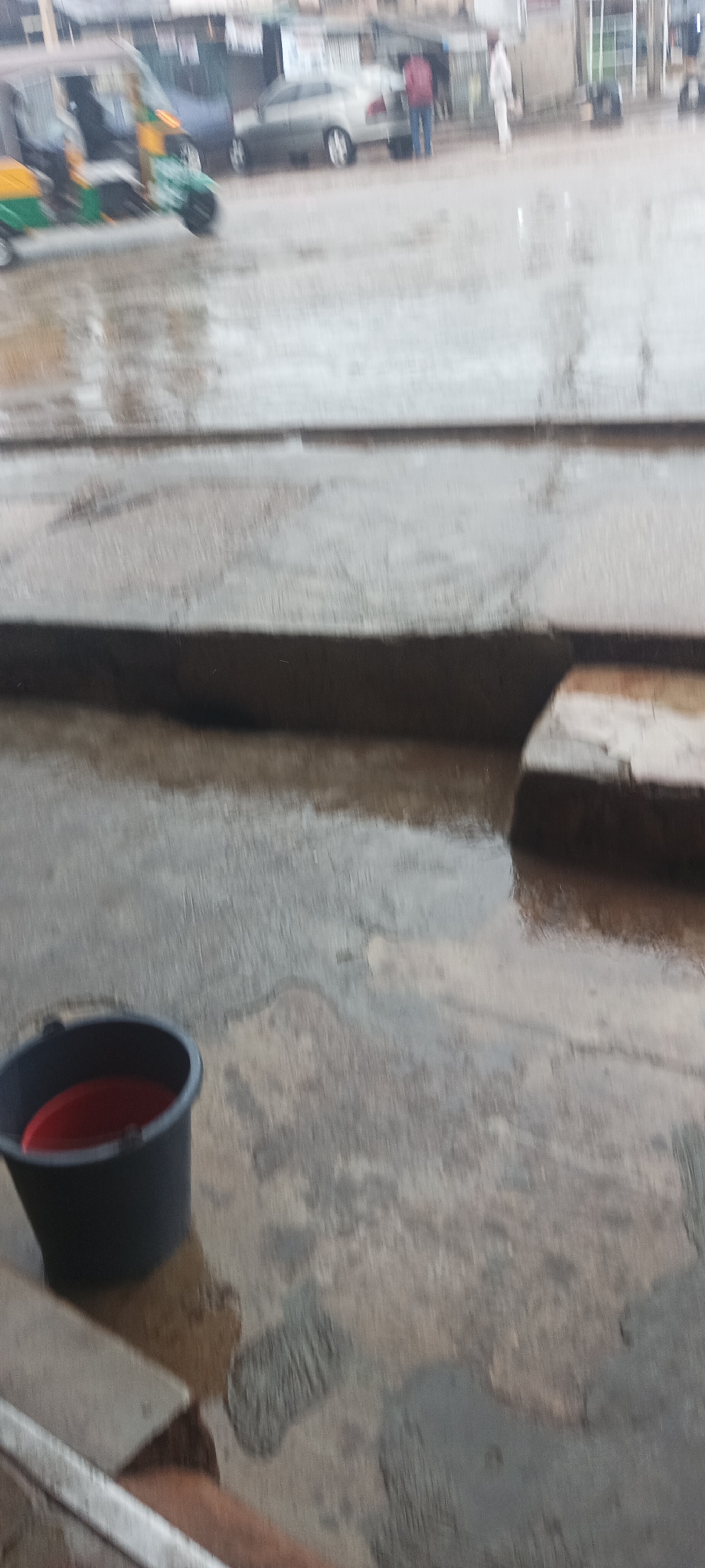 The image depicts an outdoor, street-level scene, characterized by wet concrete ground and visible puddles, suggesting recent rain or an ongoing light drizzle. In the lower-left foreground, a black bucket with a red interior sits on the damp pavement. Raised concrete structures, possibly curbs or steps, traverse the midground, creating different levels.
The background reveals a bustling street environment. On the left, a blurry yellow and green auto-rickshaw (a three-wheeled vehicle) appears to be in motion, indicating activity. Towards the center-right, a light-colored sedan car is parked or stationary. A person wearing a red top and blue pants is seen near the car's trunk, seemingly attending to it. Another person in white is partially visible further to the right. The overall setting suggests an urban or semi-urban area. The time of day is daytime, likely overcast due to the rain. No discernible text or specific emotions are evident.