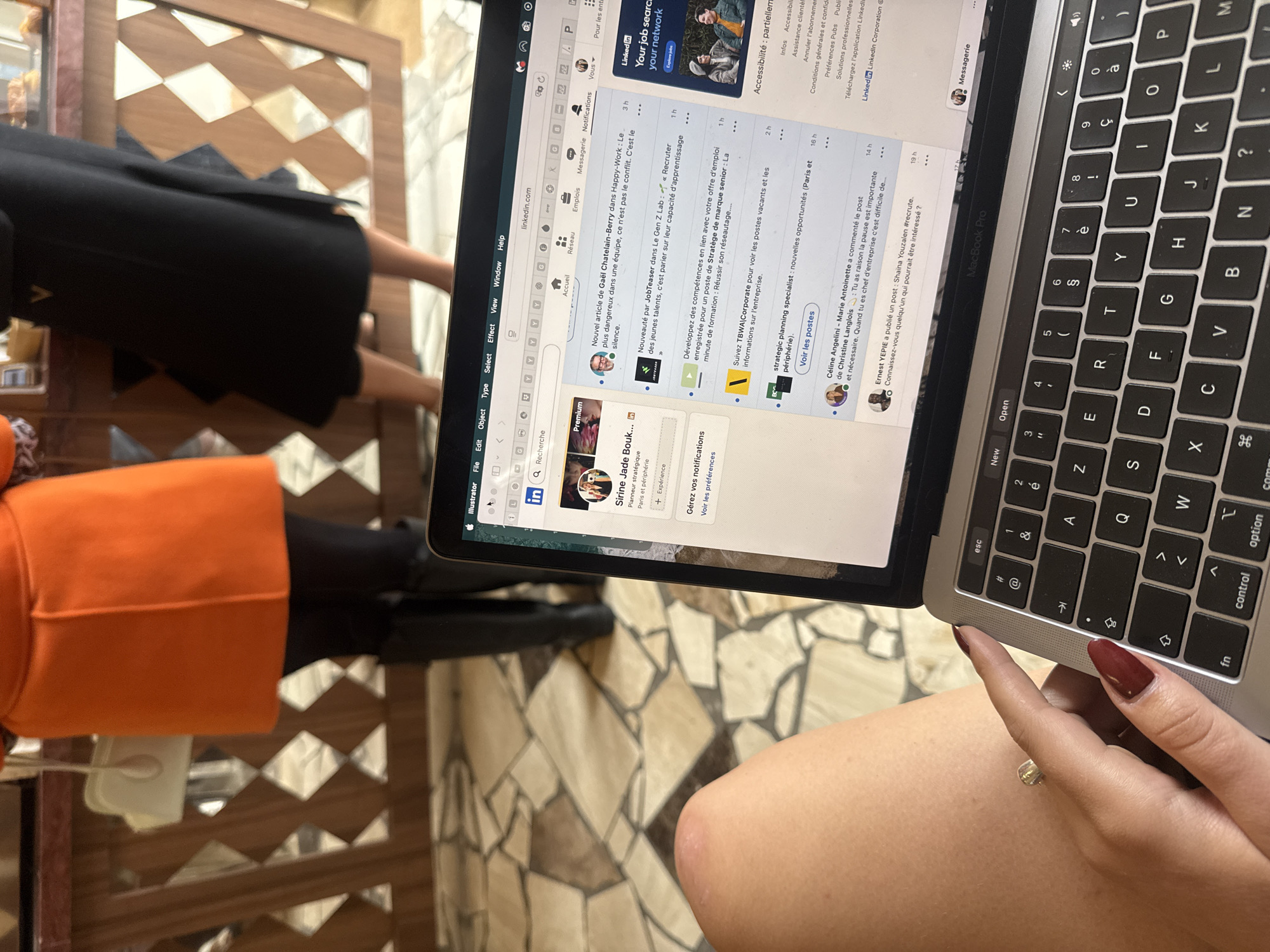 Here is a detailed description of the image:
**Scene:** The image appears to be taken from an overhead perspective, looking down at a person seated at a table while using a MacBook Pro laptop.
**People:**
* There's a person wearing an orange top and black leggings or tights.
* A second person is only partially visible, wearing a black skirt or dress.
* The primary subject has a ring on her finger and nails painted in a dark shade of red.
**Objects:**
* A MacBook Pro laptop is open, with the LinkedIn website displayed on the screen.
* The person's hand is resting on the laptop's lower right edge.
* The table has a geometric design in light brown and beige.
**Environment:**
* The location is suggested to be in Paris, France, based on the content displayed on the laptop screen (LinkedIn posts referencing Paris).
**Actions/Interactions:**
* The person is using the laptop, presumably browsing LinkedIn.
* It is possible that the laptop is resting on the person's legs.