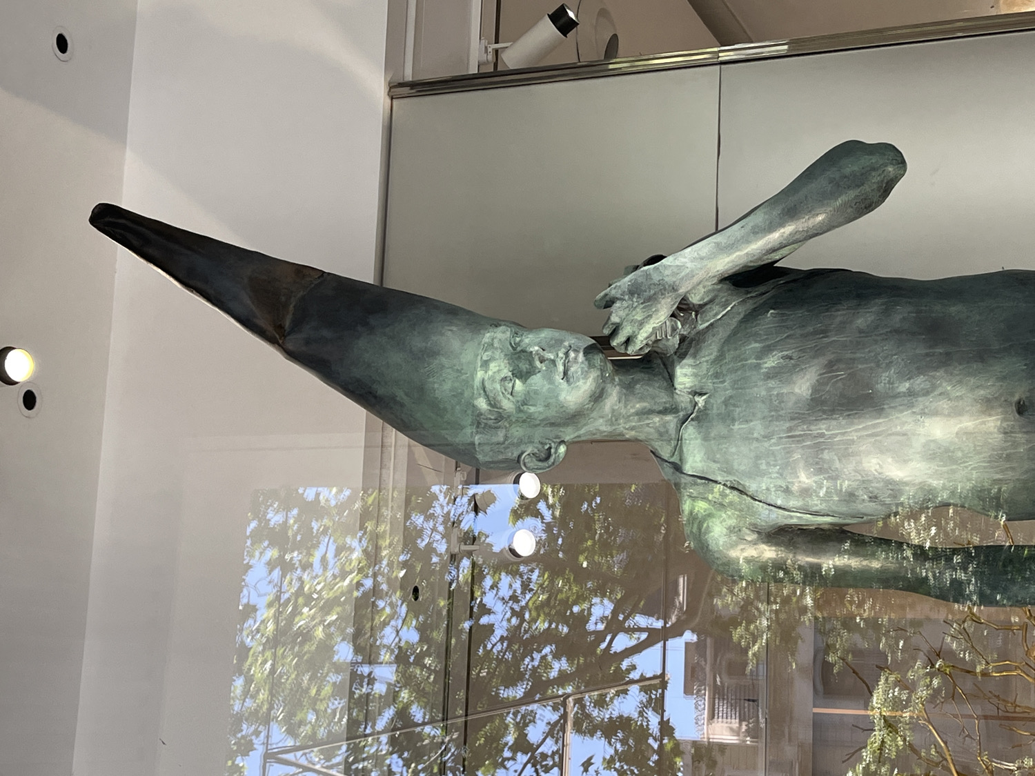 This image captures a bronze sculpture displayed within a glass enclosure, likely a shop window or museum exhibit, in Barcelona, Spain.
The primary subject is a prone or reclining human-like figure cast in bronze, exhibiting a green patina indicative of age or artistic choice. The figure has a distinctive, extremely elongated and pointed head or headwear, resembling a dunce cap or a cone, which extends upwards to the left. Its face is serene, with closed or downcast eyes, and a delicate, slightly parted mouth. One arm is bent at the elbow, with the hand gently resting near the figure's neck or lower face, creating a contemplative or relaxed pose. The body shows anatomical details such as a navel and subtle musculature.
The sculpture is lit by internal spotlights, visible as bright circular reflections on the glass, as well as ambient light. The scene is complex due to reflections on the glass panel separating the viewer from the sculpture. These reflections show a vibrant outdoor environment: lush green tree foliage, a clear blue sky, and architectural elements of a building, including balconies, suggesting an urban setting. The time of day appears to be daytime, with bright natural light.
The interaction of the sculpture with its reflected environment creates a dynamic visual effect, blurring the lines between the artwork and its setting. No discernible text is visible within the image.
