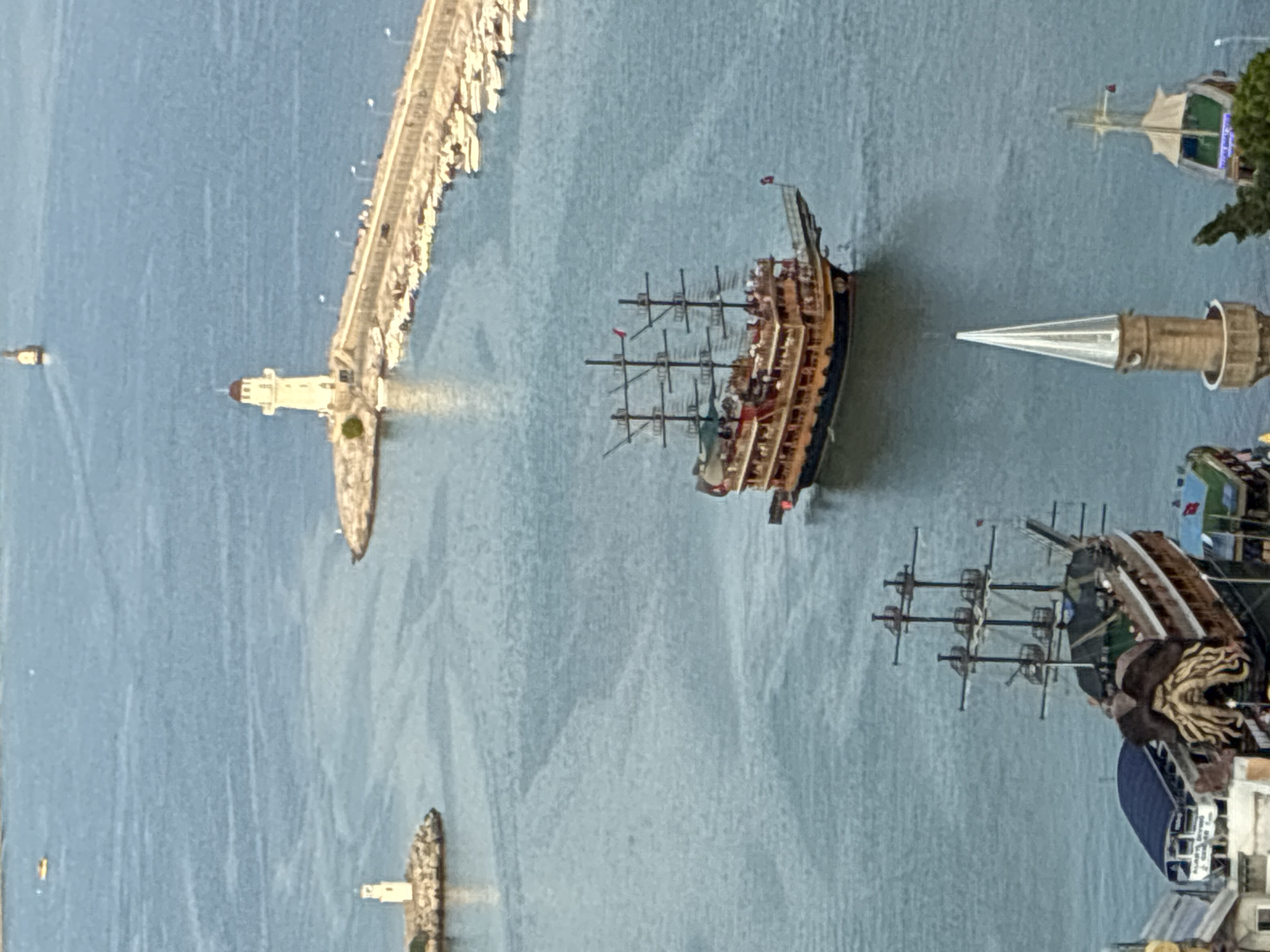 This image captures a scenic view of the harbor in Alanya, Turkey. Dominating the foreground is a large, elaborately decorated pirate-themed ship, reminiscent of a galleon, with multiple masts and decks crowded with people. To the left and slightly behind this ship, another similar but partially obscured pirate ship is docked. In the mid-ground, a long stone pier extends into the sea, lined with numerous small boats moored along its edge. At the end of this pier stands a prominent white lighthouse with a red-capped lantern room. Further in the distance, another smaller lighthouse is visible on a rocky outcrop to the left. The water is a tranquil blue, with subtle ripples and patterns indicating gentle currents. The background reveals the hazy outlines of buildings and possibly a coastline, suggesting the broader urban setting of Alanya. The sky is overcast, casting a soft, diffused light over the entire scene.