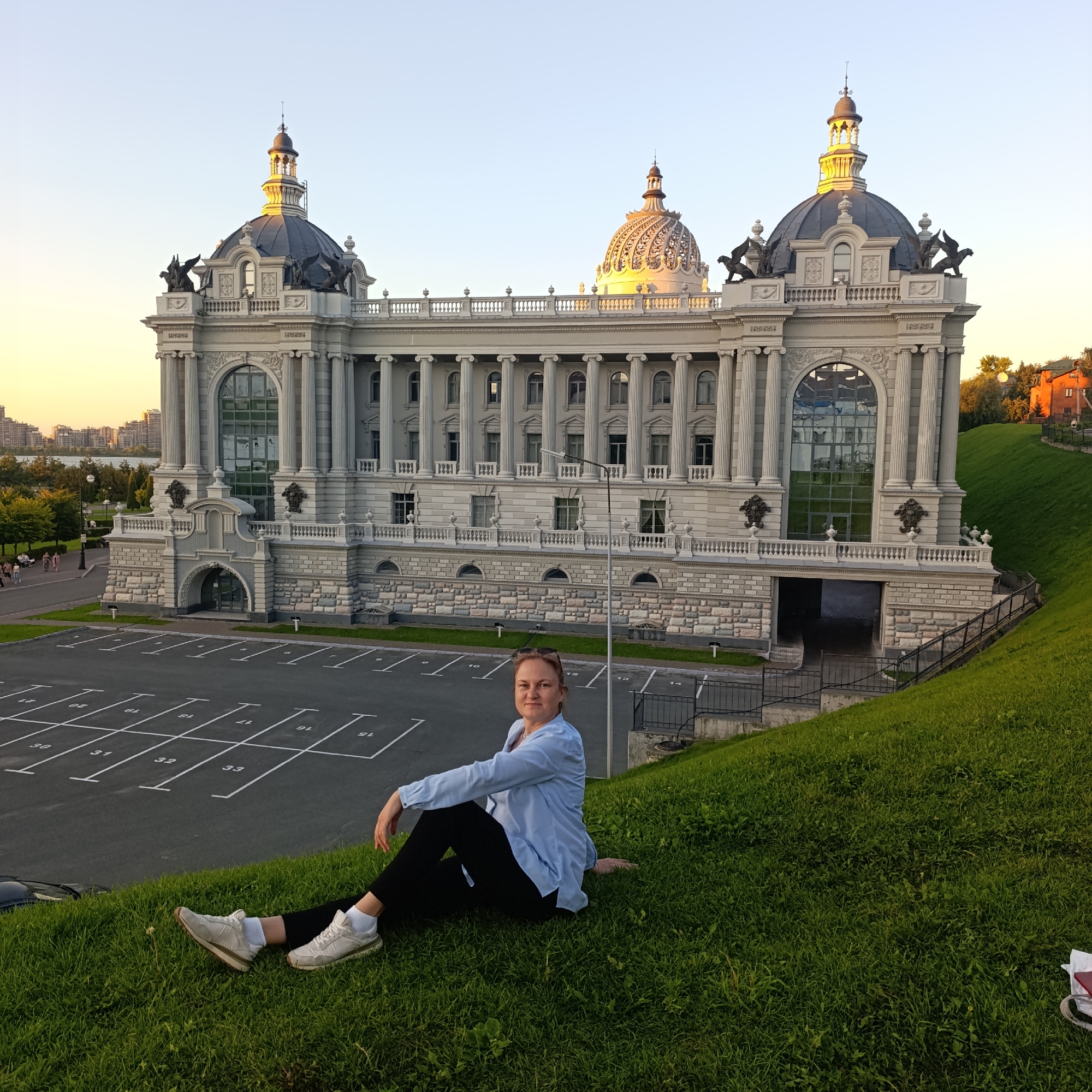 In Kazan, Russia, a woman is pictured sitting on a grassy incline facing a majestic building, likely the National Cultural Center "Kazan". The building features neoclassical architecture with prominent columns, arched windows, and multiple domes, one of which is gilded and intricately detailed. The woman, dressed in casual attire – a light blue long-sleeved shirt, black pants, and white sneakers – is looking towards the camera with a slight smile. She is positioned in the foreground, with a parking lot marked with white lines visible behind her. The scene is bathed in the warm glow of sunset, casting long shadows and highlighting the architectural details of the building. The sky is clear with a gradient of soft orange and yellow near the horizon. In the distance, the Kazan skyline and a body of water can be observed. The overall impression is a serene moment captured against a grand urban backdrop.