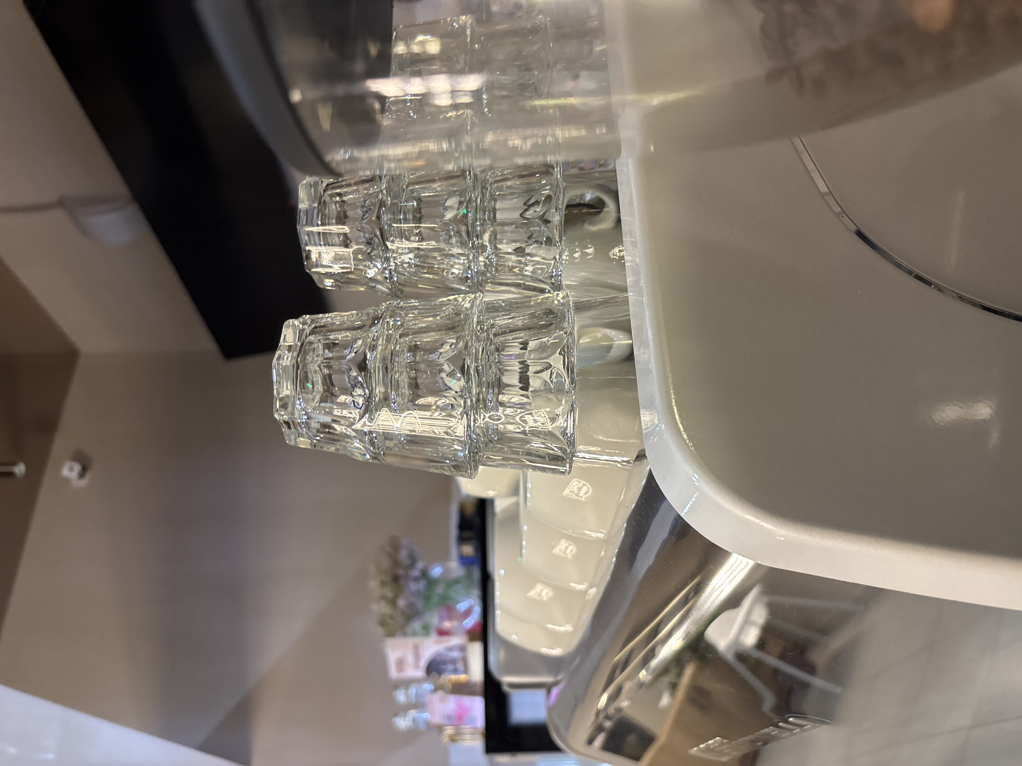 The image shows a close-up view of stacked clear glass cups on a white surface, likely a coffee machine or counter. The cups have a patterned texture. In the background, out of focus, are some bottles, a vase with flowers, and a dark shelf or structure. A glimpse of coffee beans in a container is visible on the right side of the image. The scene appears to be inside a cafe or shop. There are no people visible in the image. The lighting suggests an indoor setting with artificial lights. There is no information in the image that indicates the location is Misaylovo, Russia.
