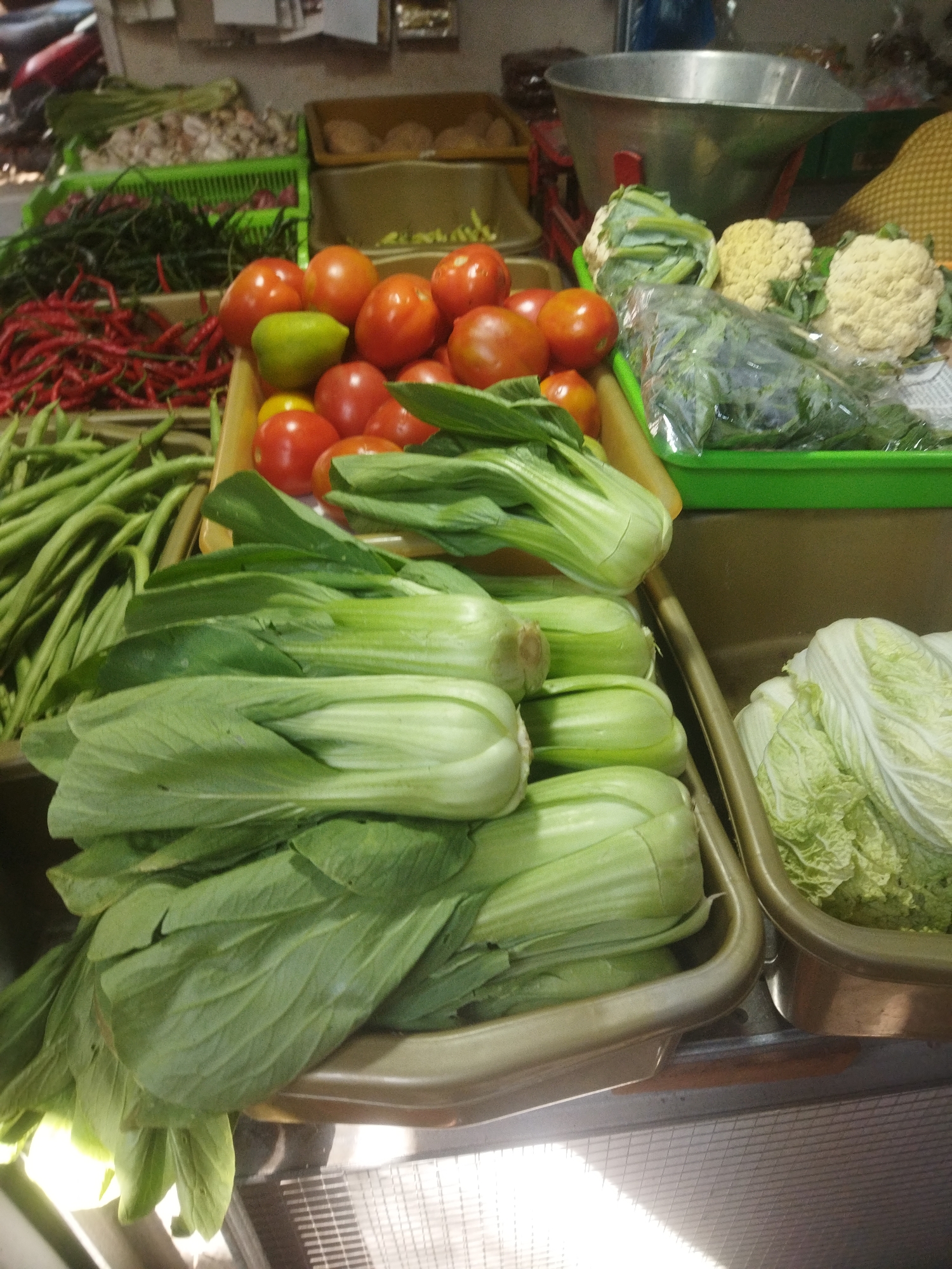 This image depicts a vibrant marketplace scene, likely in Wonogiri, Indonesia, showcasing a diverse array of fresh produce. In the foreground, several bunches of bok choy are prominently displayed in a tan-colored rectangular container. To their right, a container holds napa cabbage. Behind the bok choy, there are more leafy green vegetables, also in a tan container, with their stems visible.
Further back and to the left, a large quantity of green beans is arranged in a green plastic crate. Next to the green beans, a pile of long red chilies is also present. Above the chilies, a green crate contains more long green chilies, and behind that, there appear to be leeks or green onions.
In the center of the image, a yellow crate overflows with ripe red tomatoes. A single green pepper or lime is interspersed among the tomatoes. To the right of the tomatoes, a green plastic container holds various types of cauliflower, including white and pale yellow heads. A clear plastic bag filled with chopped green leafy vegetables is also visible in this container.
In the background, a large, metallic scale suggests active commerce. The overall environment appears to be an outdoor or semi-enclosed market, with varying levels of light and shadow suggesting natural and artificial lighting sources. No people are visible in the frame, making it a still life of market goods.