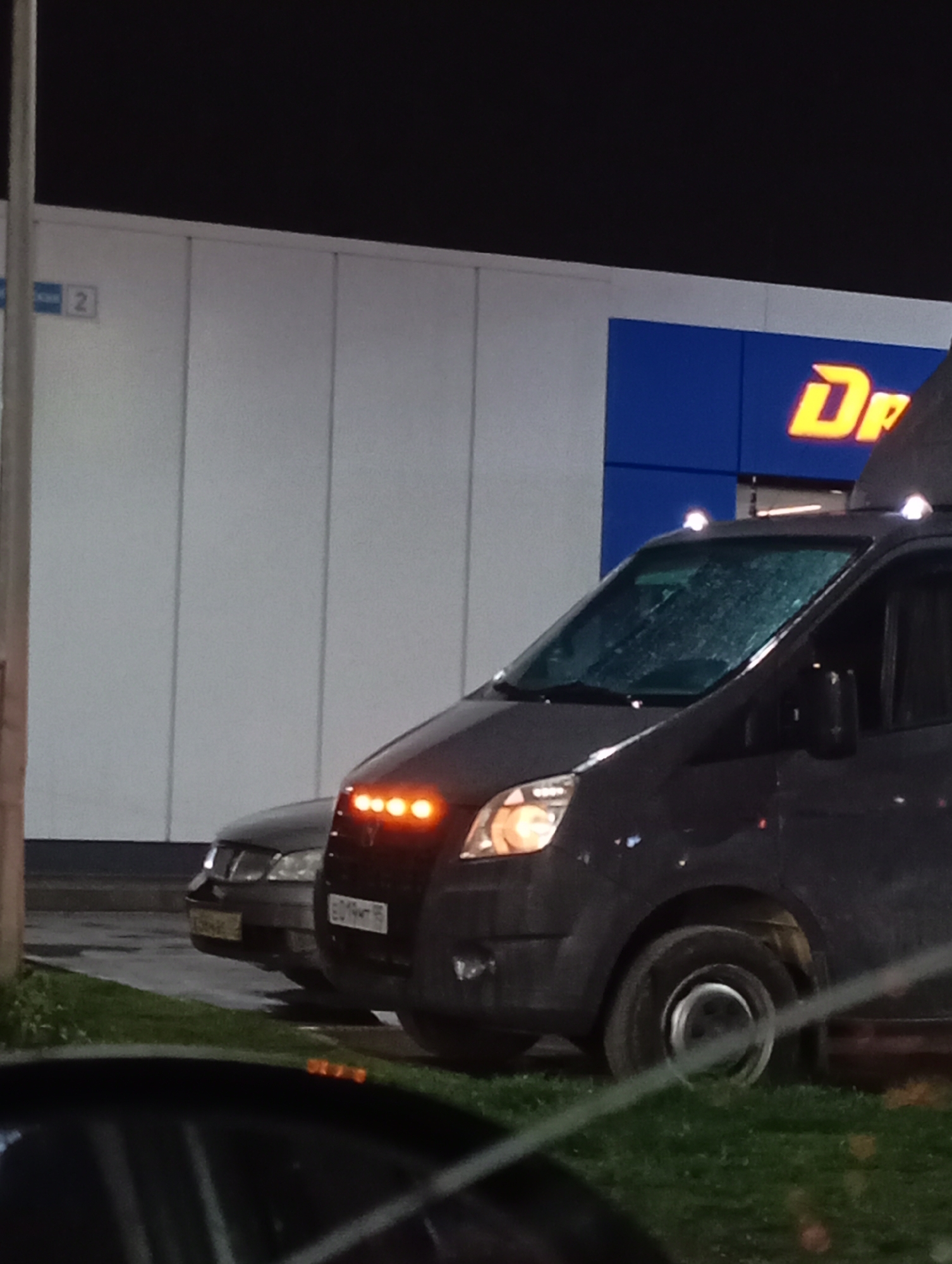 The photograph captures a nighttime scene, likely in an urban environment. In the foreground, dark grass is visible, partially obscured by what appears to be the interior of a vehicle from which the photograph was taken. Dominating the midground is a dark-colored van, parked adjacent to a sedan. The van features auxiliary lights mounted above the windshield. The sedan's license plate is partially visible. In the background, a commercial building is present. The building's facade is white, with a blue section bearing the large yellow stylized letters "Dr", likely part of a larger word. A street sign with the number "2" is also visible on the building. The cars have license plates indicating that they are from Russia. Based on the metadata, the location is in Industrialny, Russia.