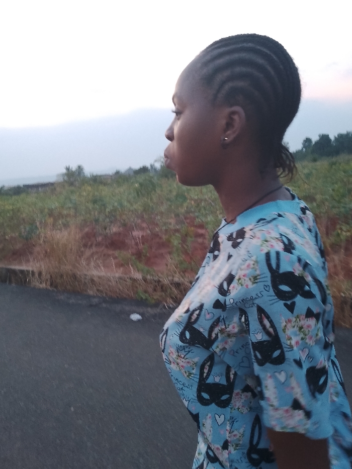 A young woman stands on a paved road. She wears her hair in braided rows, and she wears a short-sleeved light blue shirt patterned with black masks, flowers, and hearts. Her body is angled to the side, her face looking to the left. The setting is outdoors, with vegetation and distant trees visible on a slight hill in the background. The sky is light. The photograph was taken in Umueshi, Nigeria.