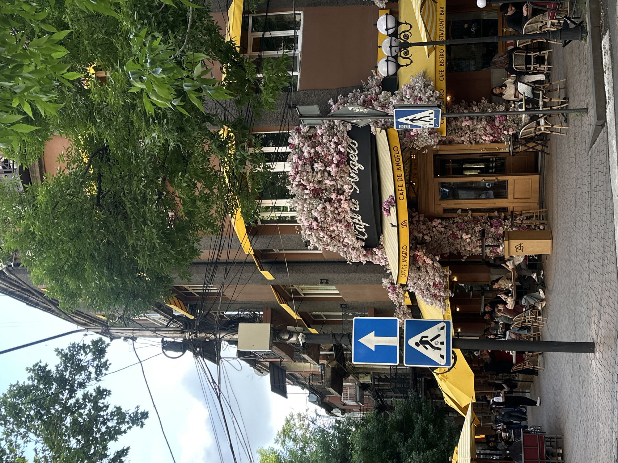 The image shows "Cafe de Angelo", a café and restaurant in Yerevan, Armenia. The building facade is decorated with flowers, and the café has a yellow awning. Diners are sitting at tables outside the restaurant. There are pedestrian crossing signs and numerous power lines running overhead. Trees partially obstruct the view.