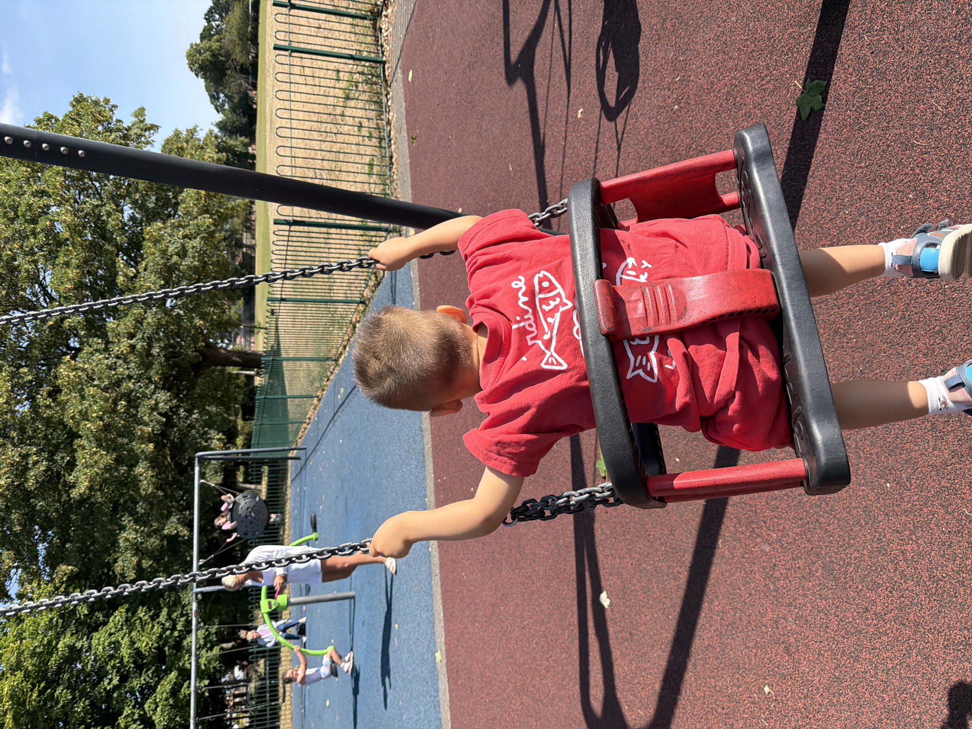 A young Caucasian boy, viewed from behind, sits on a red swing with a black seat and red frame, his right arm raised and gripping the chain. He wears a red t-shirt with a white graphic of fish and text, white socks, and blue and grey sandals. The swing is set on a red rubber playground surface, with a blue surface visible in the background. In the distance, a woman in white stands by a green swing set, while other children play on other swings, including a large spherical swing. A green metal fence encloses a grassy field with trees beyond. The scene takes place in a park, likely in Leeds, United Kingdom, under a clear blue sky.
