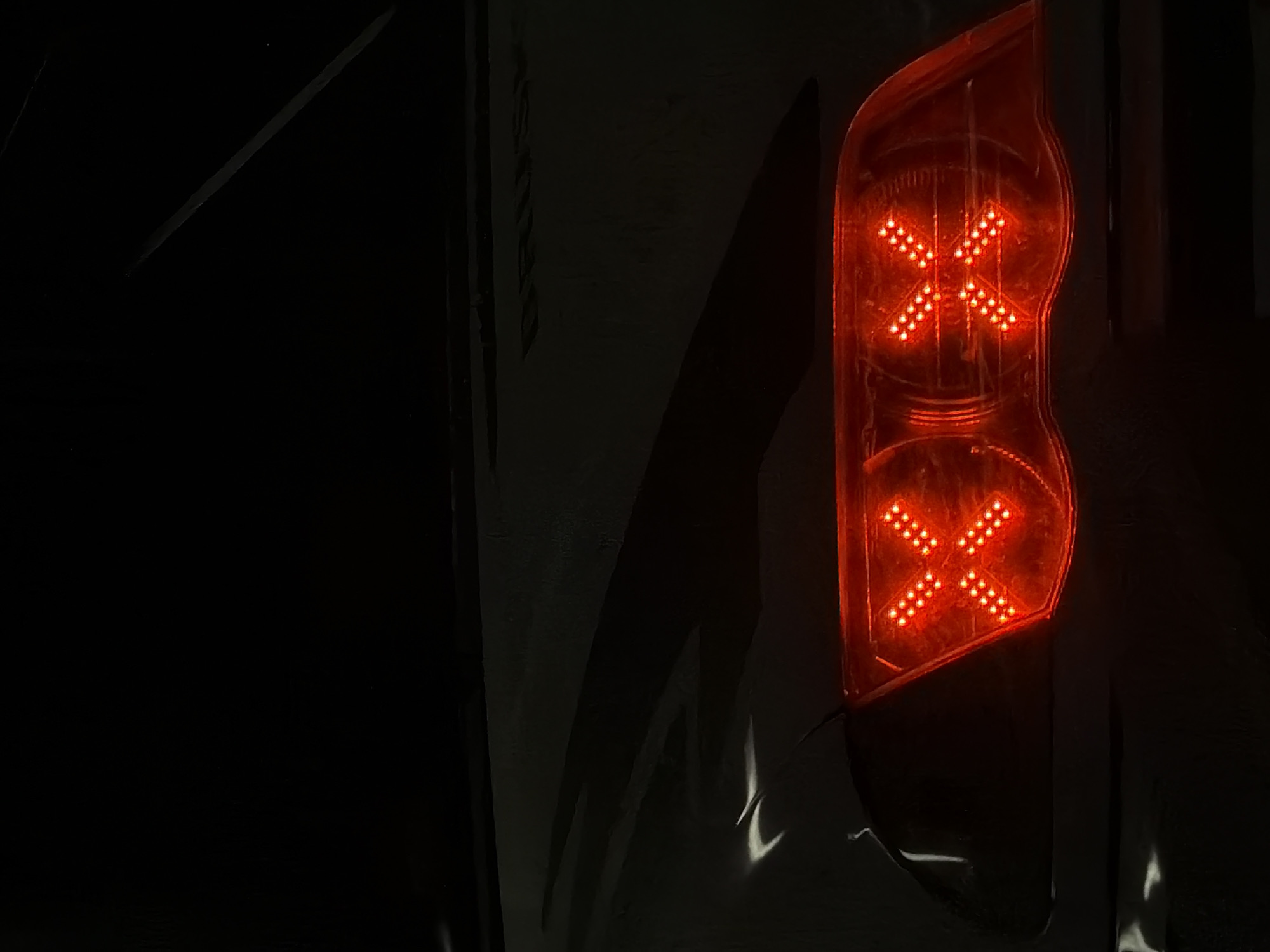 The image shows a close-up view of the rear of a car at night, with the brake lights illuminated. The brake lights are red and are designed in a distinctive "X" shape formed by multiple small LEDs. The car's bodywork is dark and appears to be wet or glossy, reflecting some ambient light. The top portion of the image is dark, showing a hint of the car's rear window or a dark sky. The word "DRUŠTVO" is visible in faint, dark lettering across the car's trunk or rear panel. There are no people or other discernable objects in the immediate vicinity. The setting is likely an outdoor urban environment, given the presence of a car and the nighttime conditions. No specific location in Plovdiv, Bulgaria can be determined from the image alone.