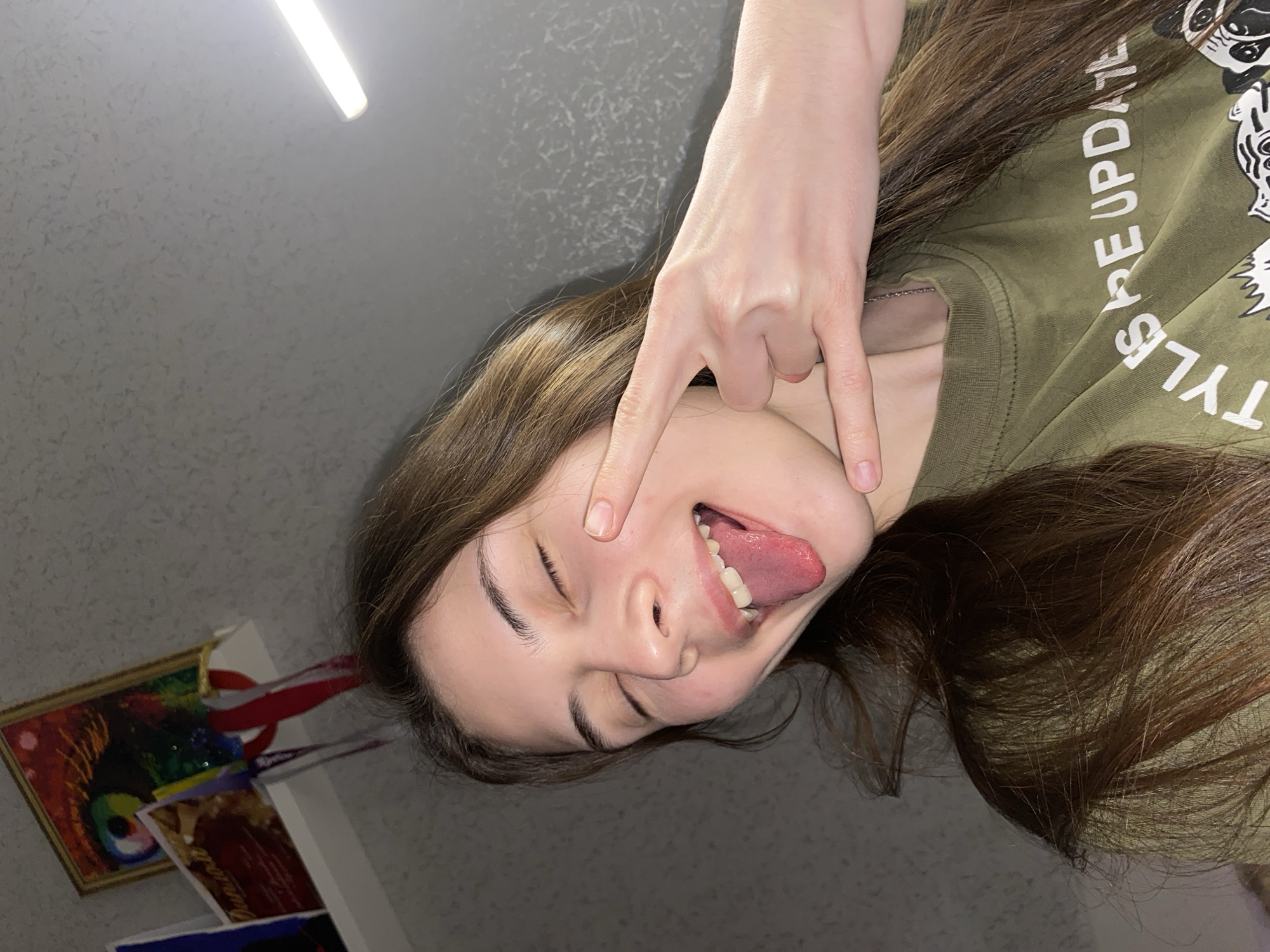 This image, captured indoors in Votkinsk, Russia, shows a young person in a playful, sideways pose. The subject, likely female, has long brown hair and is wearing a green t-shirt with the visible text "STYLE UPDATE" in white letters. She is sticking out her tongue, has her eyes closed in a mischievous expression, and is making a "rock and roll" hand gesture (devil horns) with her right hand held near her temple. Her left hand is also partially visible near her chin.
The background features a textured, light gray wall. A bright, rectangular light fixture is visible in the upper part of the frame, providing artificial illumination that suggests an indoor setting, possibly at night. In the bottom left corner, several small, framed items or cards are partially visible, including one with vibrant, abstract artwork. The overall mood is energetic and expressive.