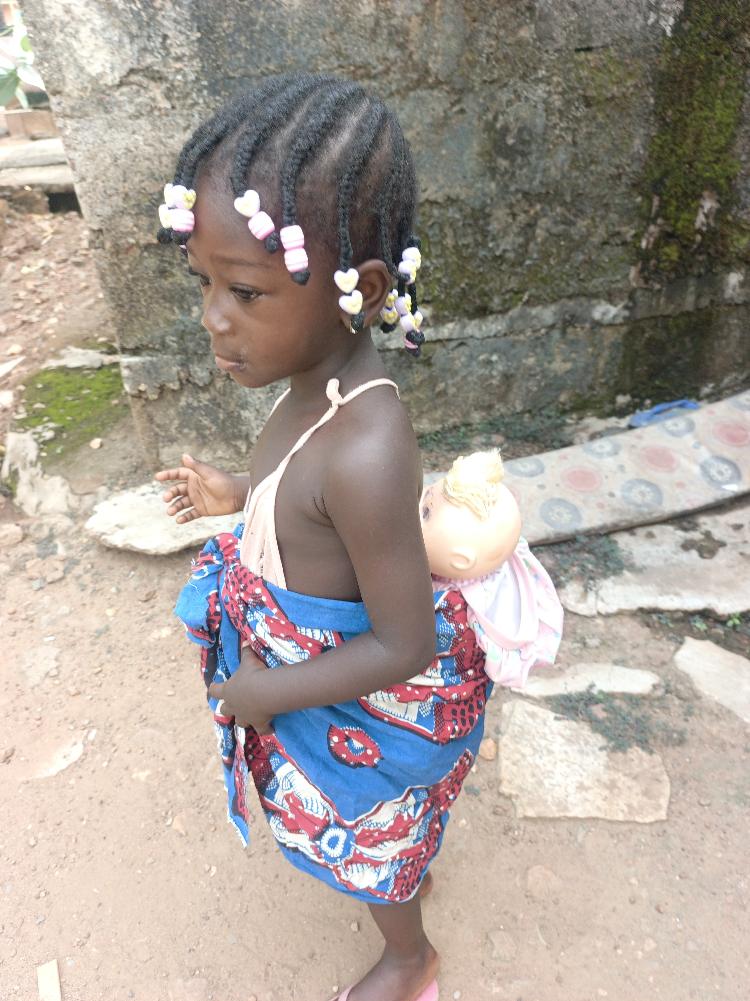 A medium shot shows a young Black girl standing outdoors in Akwanga, Nigeria, holding a doll wrapped in fabric on her back. The girl faces slightly to her left, looking downwards with a contemplative expression. Her dark hair is styled in numerous small braids, each adorned with colorful heart-shaped beads in pink and yellow. She wears a light-colored, possibly peach or beige, strappy top.
Around her waist and back, a vibrant blue fabric with distinctive red and white patterns is tied, securely holding a doll. The doll, partially visible on her back, has blonde hair and is dressed in a light pink garment, mimicking a baby being carried. The child is wearing small pink flip-flops on her feet.
The setting features an unpaved ground of reddish-brown dirt or sand, with scattered small stones and patches of sparse greenery. In the background, there is a rough, weathered concrete wall, partially covered with moss and lichen. To the right, a patterned mat or piece of fabric lies on the ground. The natural lighting suggests it is daytime, with soft, indirect illumination. No text is visible in the image.