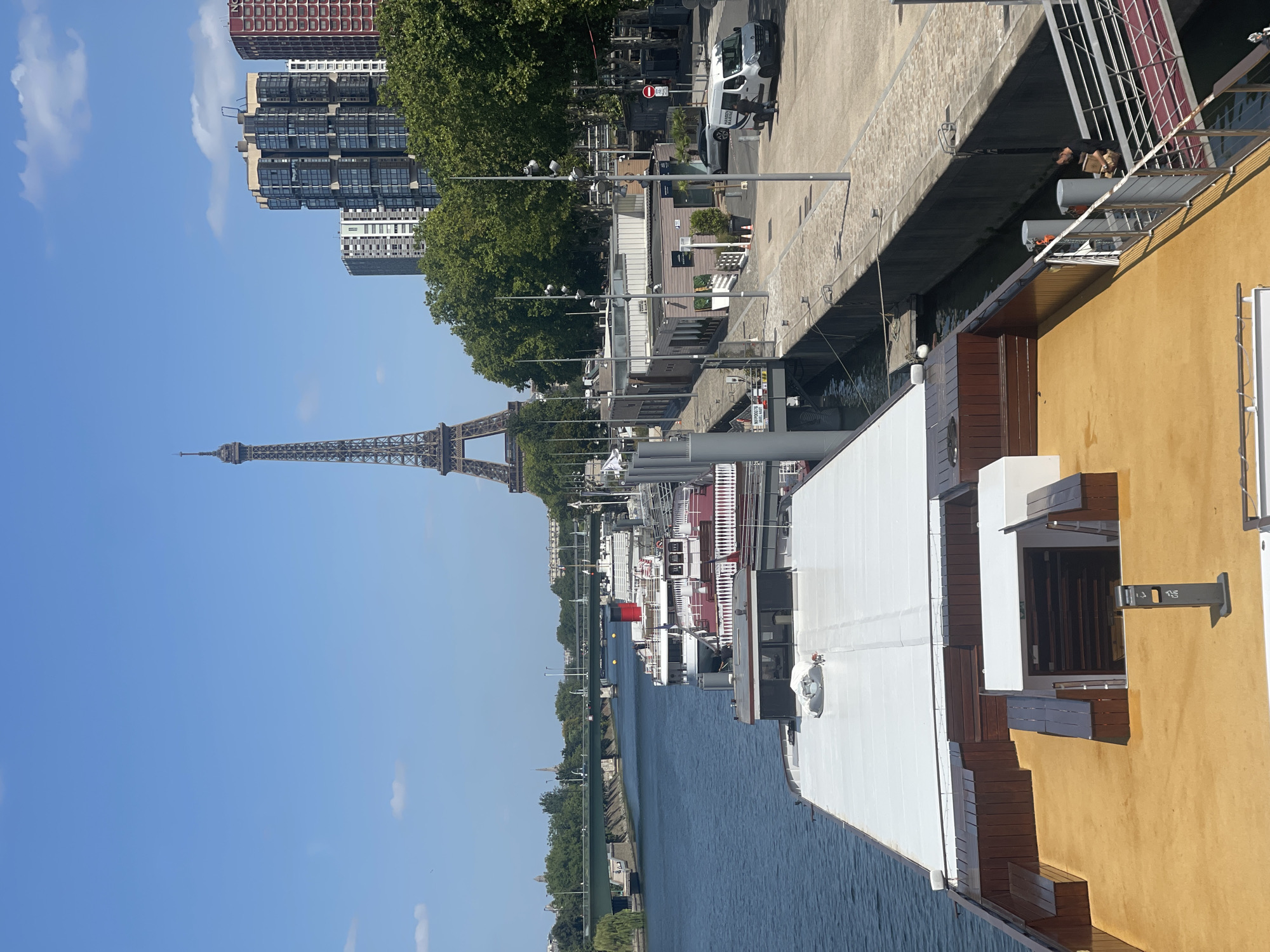 The image shows a view of the Seine River in Paris, France, with several houseboats docked along the embankment. In the background, the Eiffel Tower is visible, along with modern buildings and trees. The foreground features the deck of a houseboat, with its white rooftop and a yellow-painted area with wooden benches. A man is seen on the dock, carrying boxes and walking towards a ramp leading onto another boat. A white van is parked nearby on the street. The sky is clear and blue, indicating a sunny day.