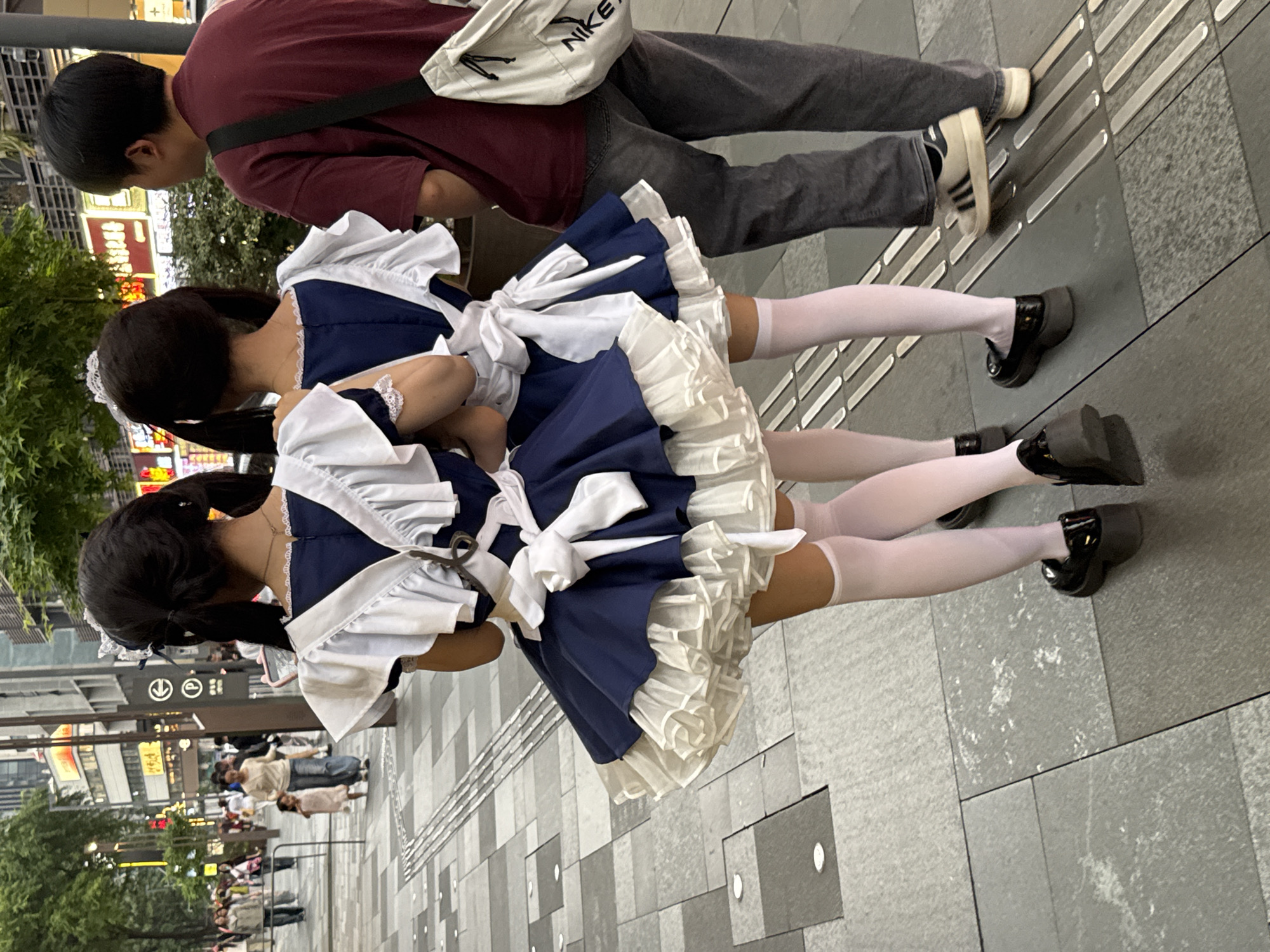 Two women dressed in matching maid outfits walk on a paved street alongside a man in a red shirt. The women's maid outfits are blue and white with frilly aprons and white thigh-high socks. They wear black Mary Jane shoes. The man wears a red shirt, gray pants, and black and white sneakers, and he carries a white bag with black straps. The street has dark gray and light gray paving stones, and a line of metal bumps for the visually impaired is visible. Buildings and trees are visible in the background, suggesting an urban setting in Jinjiang District, China. People walk along the street in the distance.