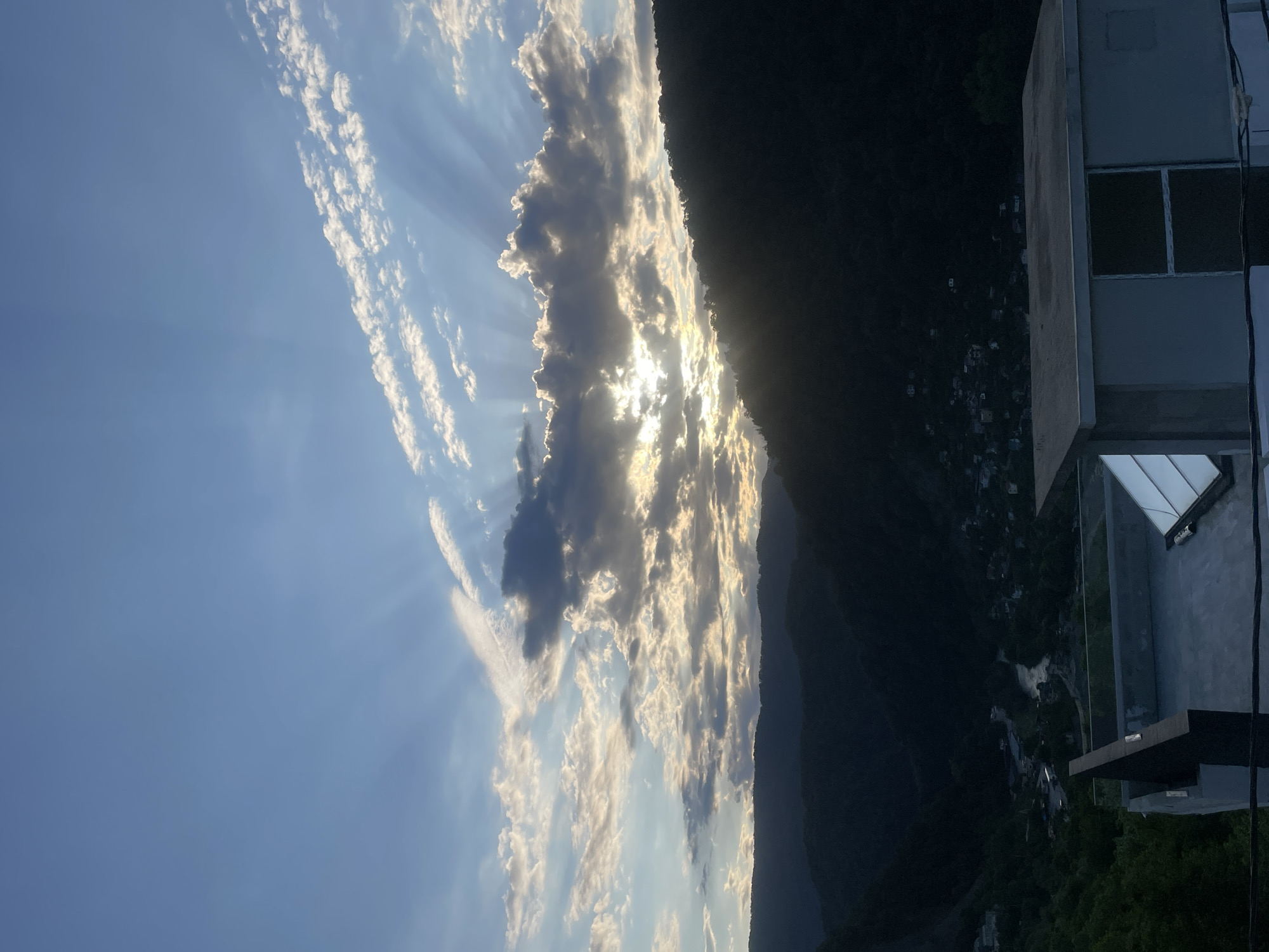 A scenic outdoor photograph captures a dramatic sky unfolding over a mountainous landscape, with a modern building situated on the right. The upper portion of the image is dominated by a bright blue sky, partially adorned with elongated bands of white and grey clouds. The sun, positioned low in the sky slightly right of center, is mostly obscured by a thick bank of clouds, from which striking golden crepuscular rays fan out, illuminating the cloud edges and creating a breathtaking visual effect.
The lower and left sections of the image reveal dark, silhouetted mountains and hills, cast in deep shadow or twilight, hinting at the vastness of the natural terrain. Some patches of green foliage are faintly visible among the dark forms. On the far right, a contemporary building, constructed from light-colored concrete or stone with a flat roof and large, dark-paned windows, stands overlooking the vista. Scattered faint lights in the distant valley or on the mountain slopes suggest human habitation within the vast landscape.
The prominent sunbeams and deep shadows indicate that the photograph was taken during either sunrise or sunset. The overall mood is one of awe-inspiring natural beauty and tranquility. No visible text is present in the image. The scene is consistent with a mountainous region such as Baranovka, Russia.