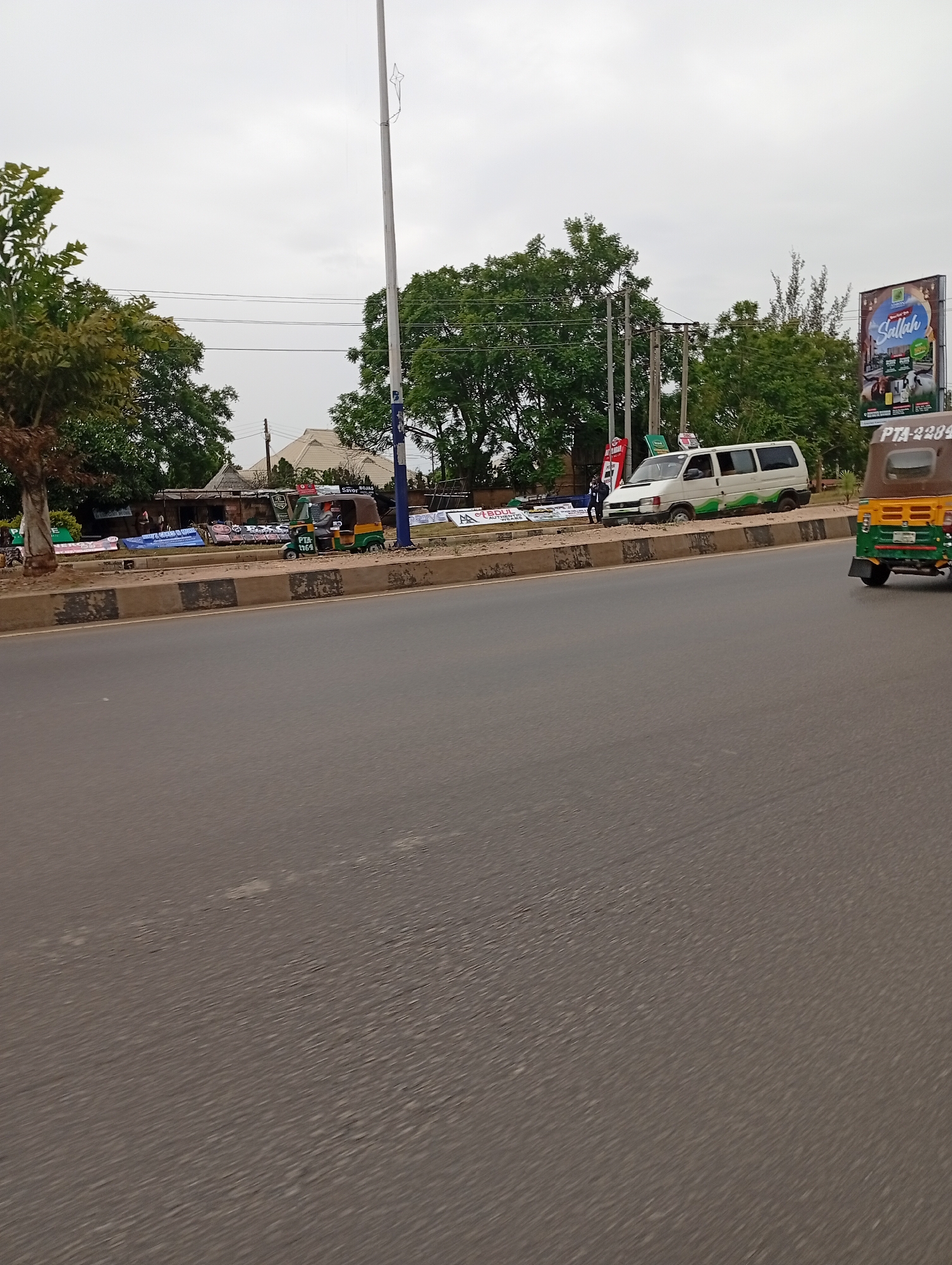 A street scene in Jos, Nigeria, is depicted on an overcast day. The foreground is dominated by a paved road with a faint pattern of wear. The middle ground features a raised concrete barrier separating the road from a sidewalk lined with various signs and a few small structures, including what appear to be market stalls or shops. Three-wheeled vehicles (Keke Marwa) and a white van are parked or moving along the side of the road. The background consists of trees, utility poles, and a billboard advertising "Sallah". The overall impression is of a bustling urban environment.