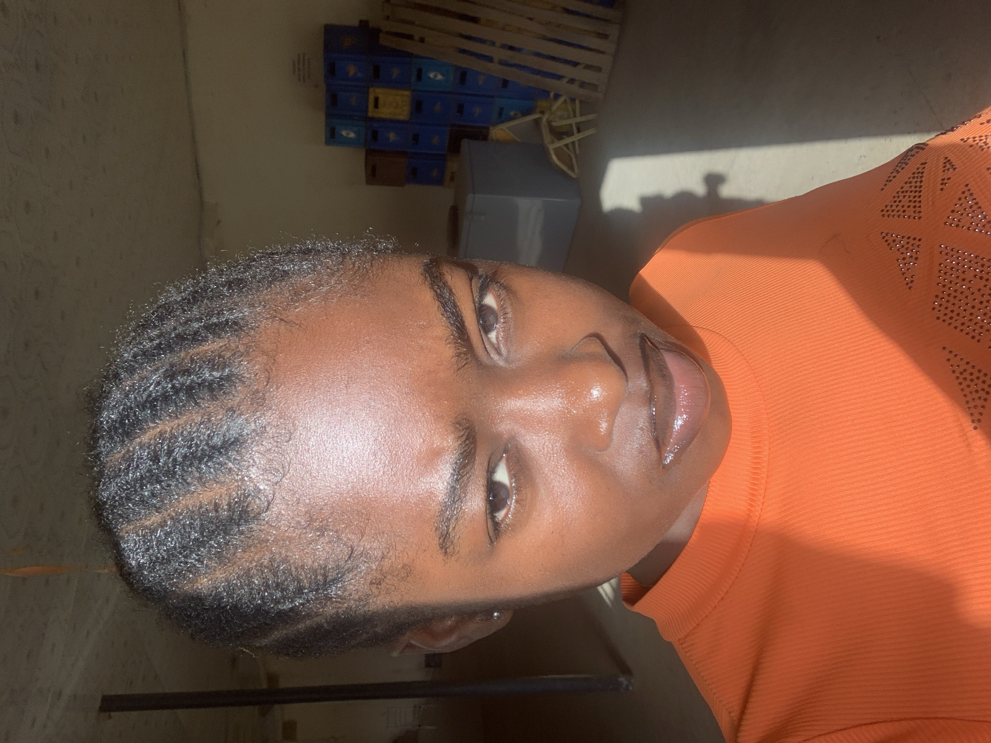 A close-up portrait of a Black woman with braided hair wearing an orange shirt. The background includes stacked blue and yellow crates and a grey box. The location is likely indoors in New Stadium, Nigeria.